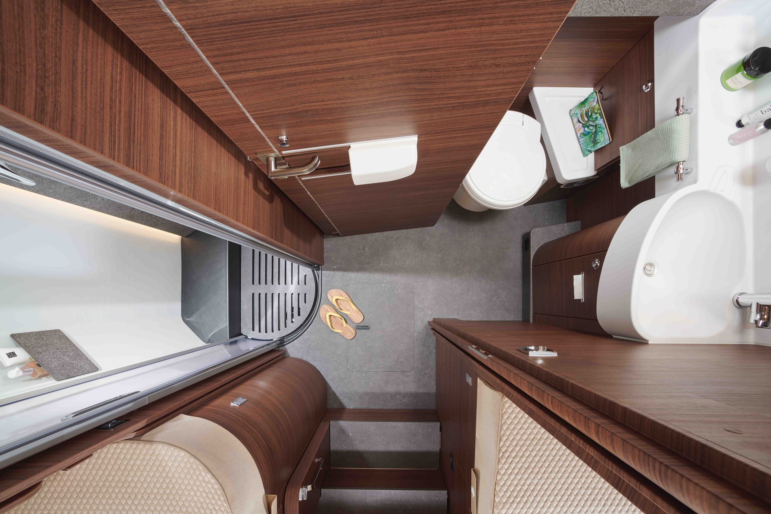 Ein kompaktes Wohnmobil-Badezimmer von oben gesehen, mit Dusche, Toilette, Waschbecken, Holzschränken, einem Handtuch, Toilettenartikeln und einem Paar gelber Sandalen auf dem grauen Boden.