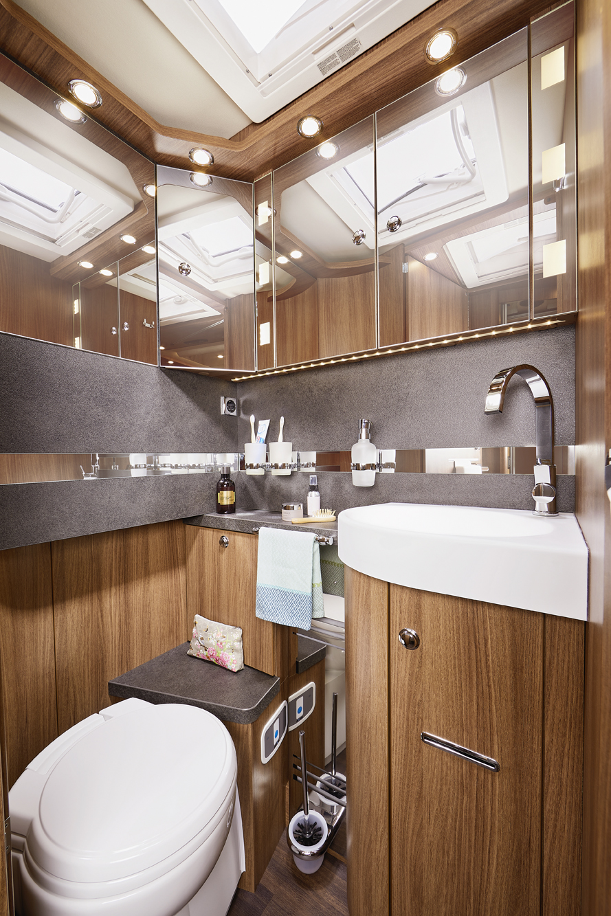 Modernes Wohnmobil-Badezimmer mit holzgetäfelten Schränken, verspiegelten Wänden, einer weißen Toilette, einem Waschbecken mit verchromtem Wasserhahn, Toilettenartikeln, Handtüchern und Deckenbeleuchtung. Oberlichter über dem Bad lassen natürliches Licht herein.