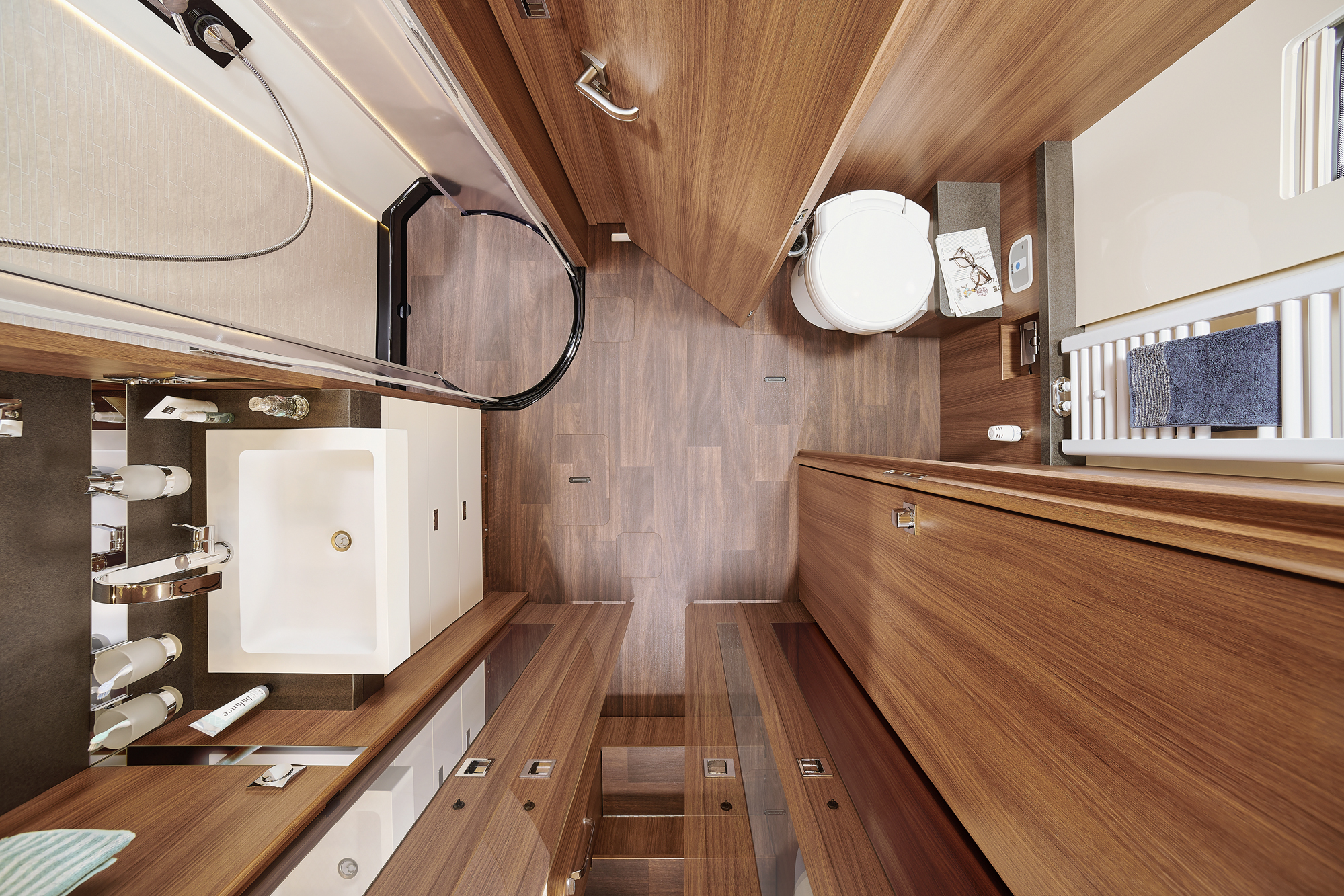 Ein modernes Wohnmobil-Badezimmer mit holzvertäfelten Wänden und Böden, einer Dusche mit Glastür, einem weißen Waschbecken und einer Arbeitsplatte, einer Toilette und Ablageschränken, von oben gesehen.