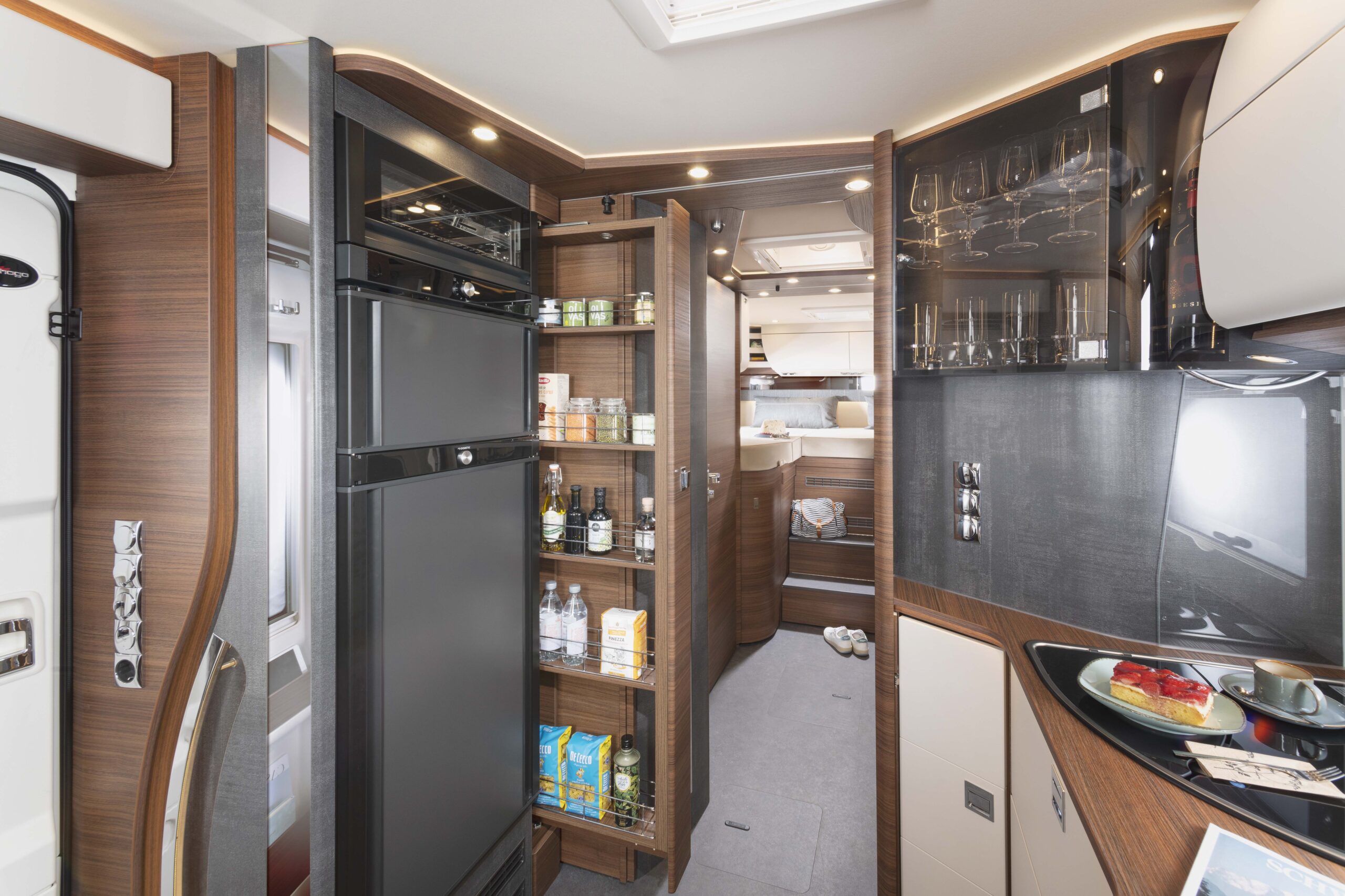 Moderne Wohnmobilküche mit Holzschränken, einem schwarzen Kühlschrank, offenen Vorratsregalen mit Lebensmitteln und Getränken, einem Glasschrank, einer Spüle und einer Theke mit einem Teller mit Lebensmitteln. Im Hintergrund ist der Schlafbereich zu sehen.