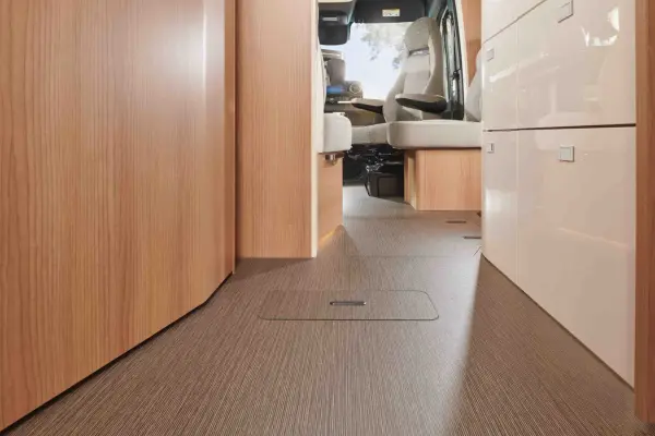 Blick vom Boden eines Carthago C Tourer Teilintegrierten Wohnmobils, der helle Holzschränke, Schubladen und beige gepolsterte Sitze zeigt, während das Sonnenlicht durch die Fenster in der Nähe des vorderen Fahrbereichs einfällt.