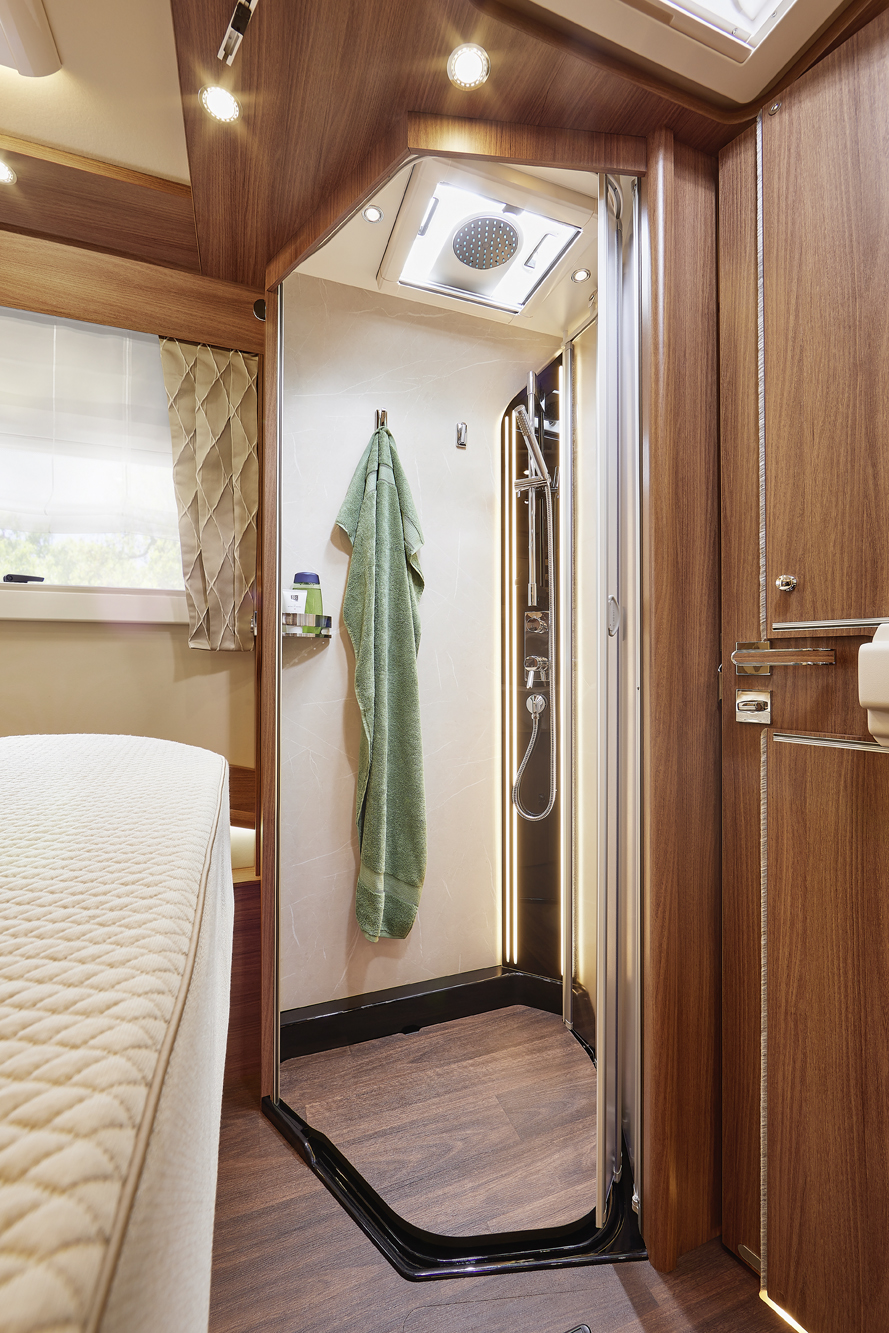 A compact shower with wood-paneled walls and flooring inside a motorhome. The shower has a glass door, a green towel hanging on a hook, and a built-in showerhead with toiletries on a corner shelf.