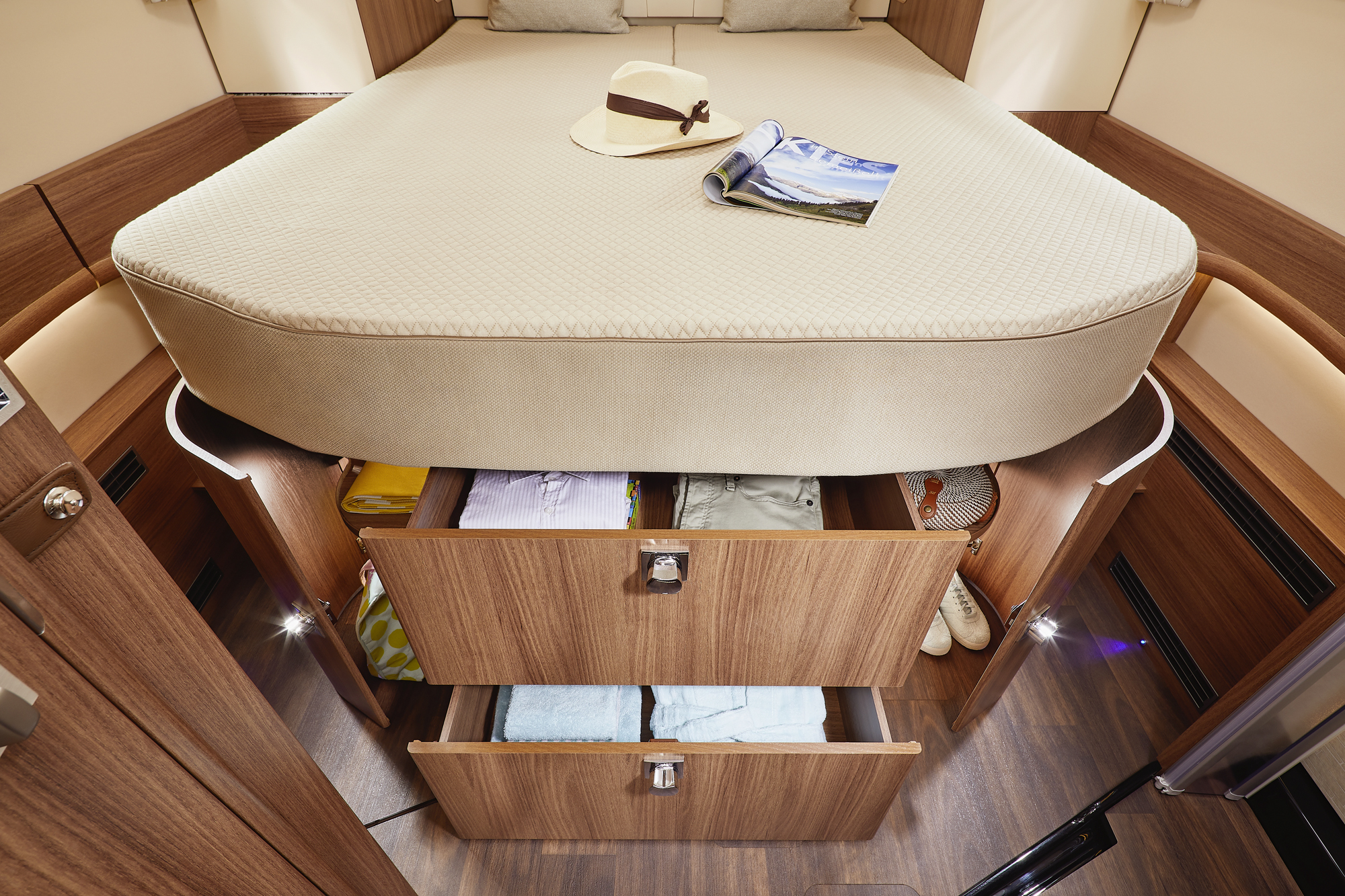 Een keurig opgemaakt bed met een hoed en een tijdschrift erop. Onder het bed zijn twee open houten lades gevuld met opgevouwen kleding en schoenen, ingebouwd in een compact, modern houten opbergmeubel.