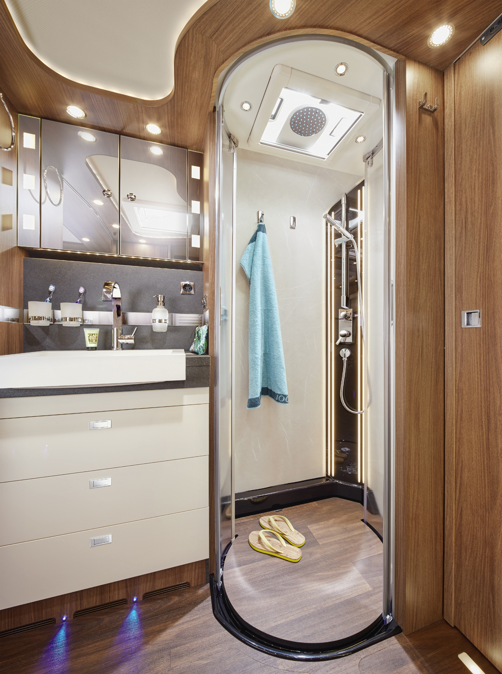 Modernes Wohnmobil-Badezimmer mit Holzvertäfelung, gebogener Glastür, begehbarer Dusche, Regenduschkopf und innen hängendem Handtuch. Auf dem Boden liegen beigefarbene Flip-Flops. Waschbecken, Spiegel und Schubladen sind auf der linken Seite sichtbar.