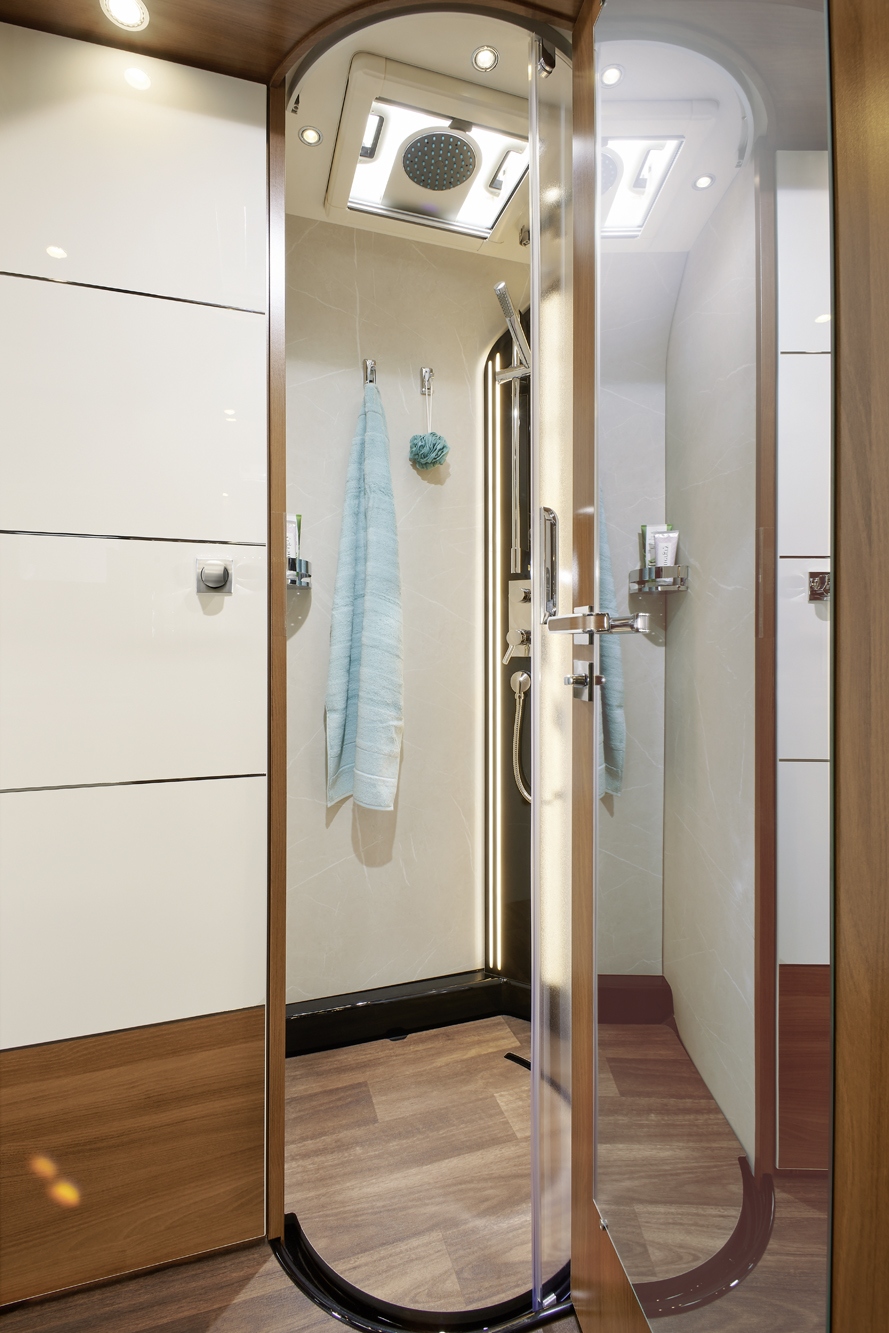 Eine moderne Dusche mit einer gewölbten Glastür, hellbraunen Fliesenwänden, Holzfußboden, einem Regenduschkopf und zwei hellblauen Handtüchern an der Rückwand. In den Regalen sind Toilettenartikel untergebracht, und Deckenleuchten erhellen den Raum.