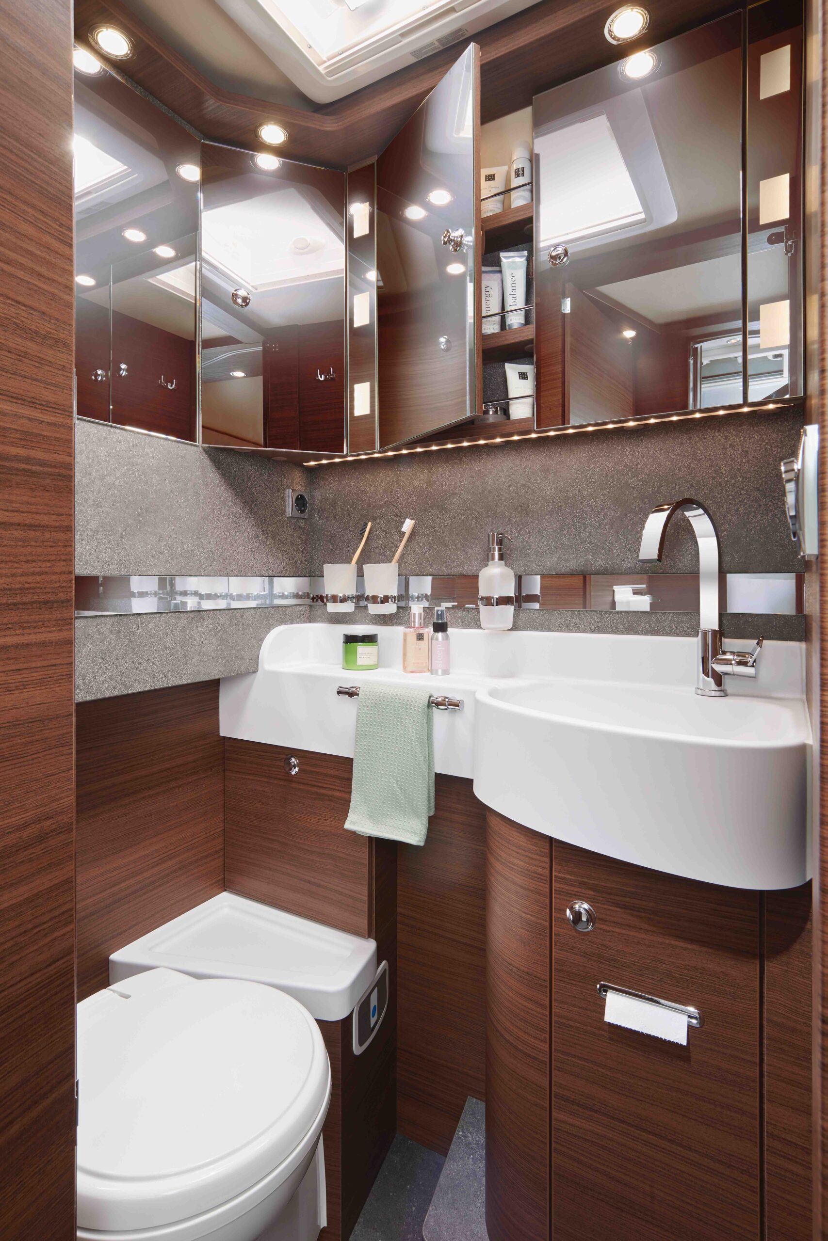 Modernes Badezimmer mit holzgetäfelten Schränken, weißem Waschbecken, silbernem Wasserhahn und einer Toilette. Ein grünes Handtuch hängt über dem Waschbecken, und Toilettenartikel, Zahnbürsten und Spiegel sind ordentlich im Raum verteilt.