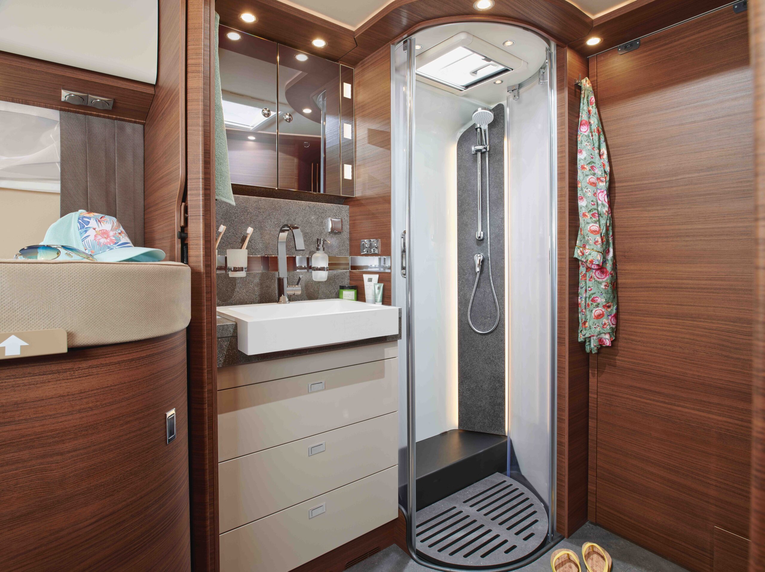 Modernes Wohnmobil-Badezimmer mit Holzvertäfelung, weißem Waschbecken und Schubladen, Dusche mit Glastür, Bademantel an einem Haken, Sonnenhut und Sonnenbrille auf einem Regal und Hausschuhe auf dem Boden. Die helle Beleuchtung schafft eine gemütliche Atmosphäre.