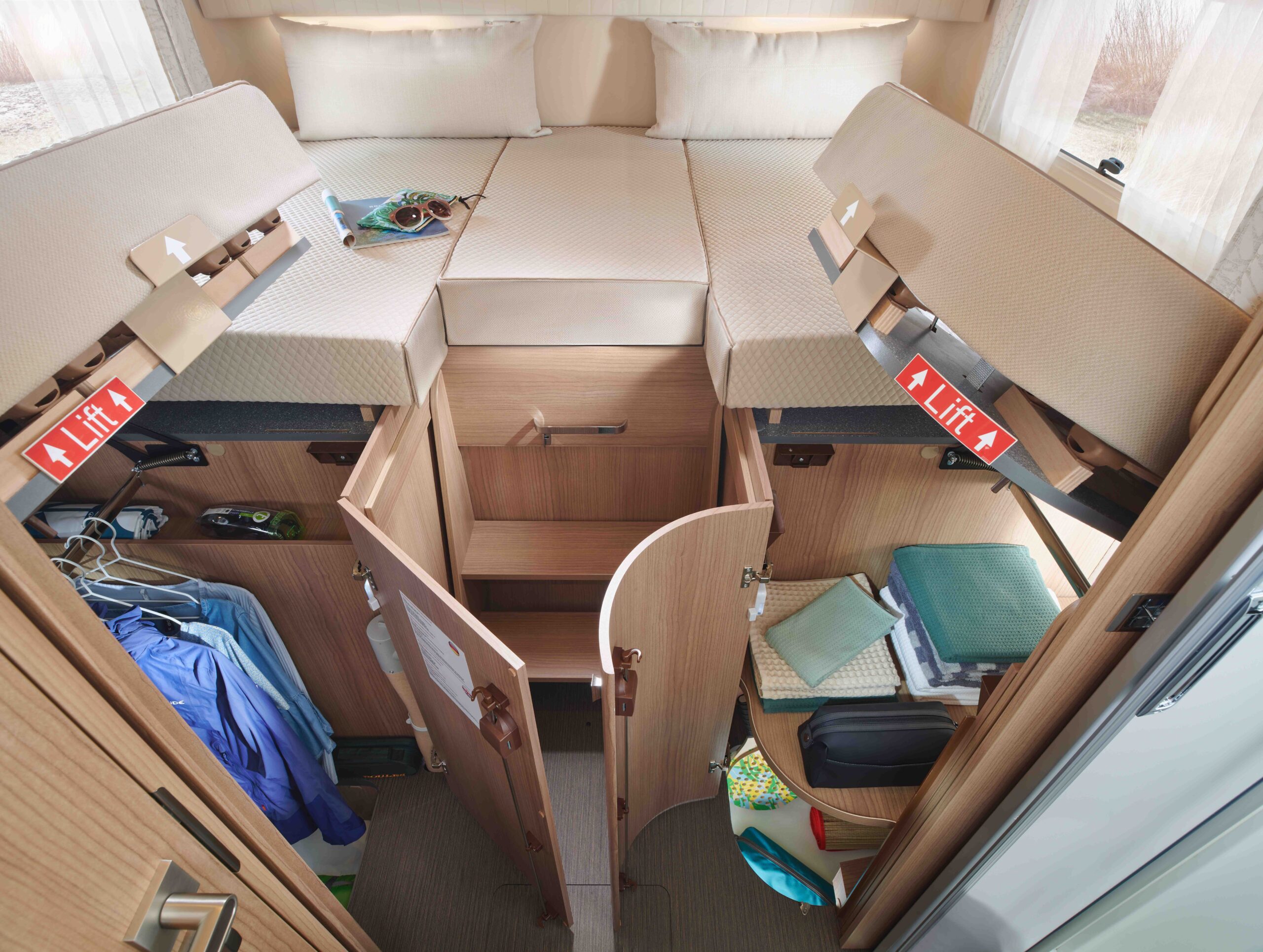 In den offenen Staufächern unter den Einzelbetten eines Wohnmobils finden Kleidung, Schuhe, Bücher und Reiseutensilien Platz. Zwei "LIFT"-Aufkleber sind an den Unterbettfächern zu sehen. Licht strömt durch verdunkelte Fenster herein.