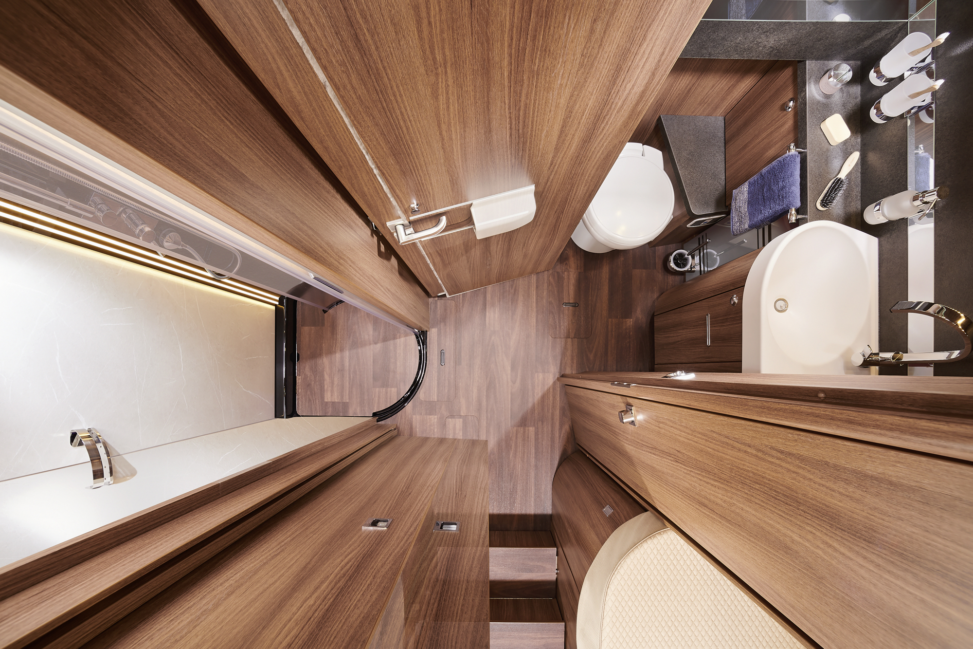 Ein modernes Wohnmobil-Badezimmer, von oben fotografiert, mit holzgetäfelten Wänden und Schränken, einer weißen Toilette, einem Waschbecken, einer Badewanne und einem Spiegel mit Toilettenartikeln und Handtüchern.