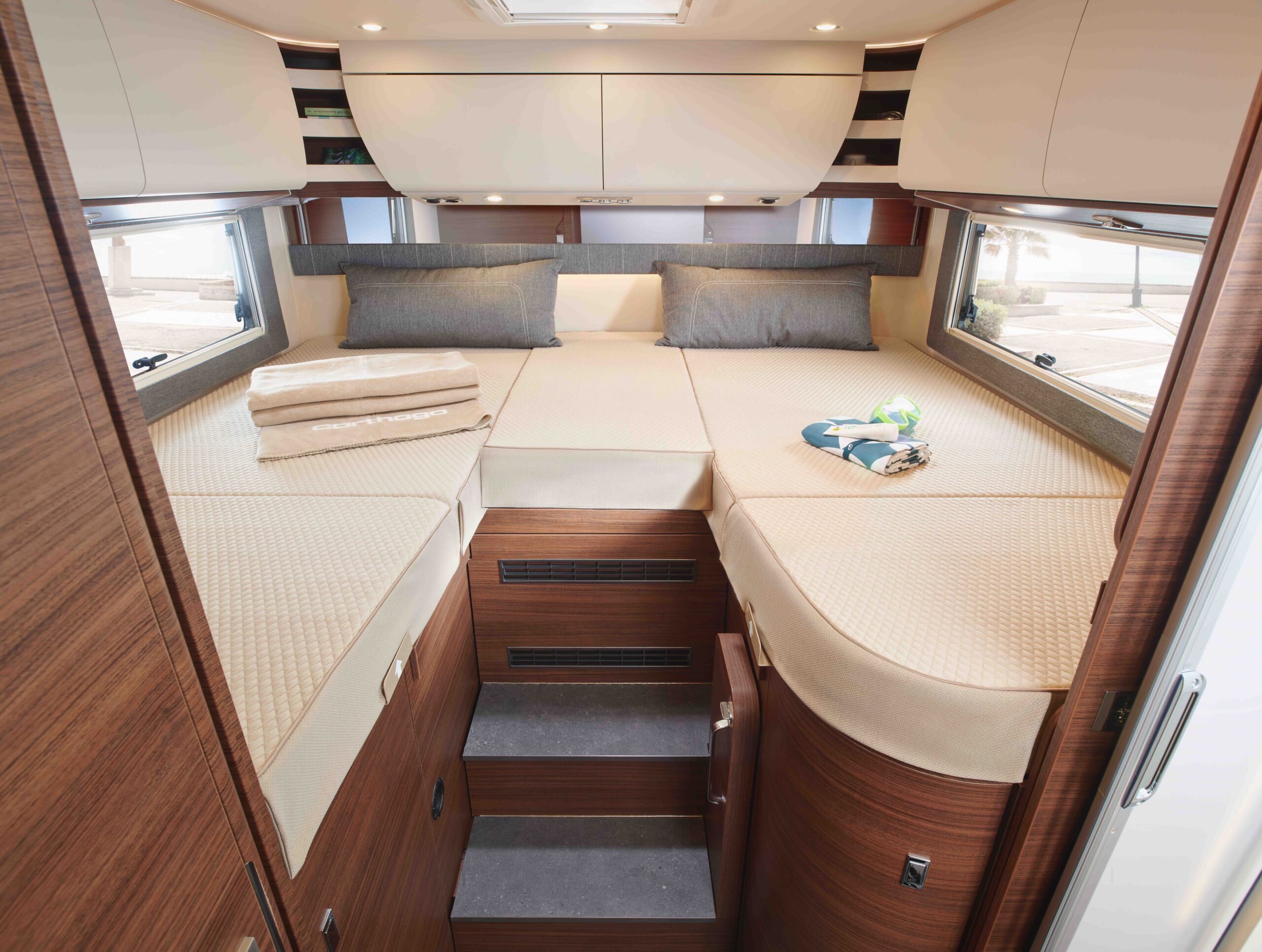 Ein gemütliches Wohnmobil-Interieur mit Einzelbetten auf beiden Seiten, hellbrauner Bettwäsche, grauen Kissen, einer Treppe in der Mitte, Holzschränken, gefalteten Handtüchern und großen Fenstern, die natürliches Licht hereinlassen.