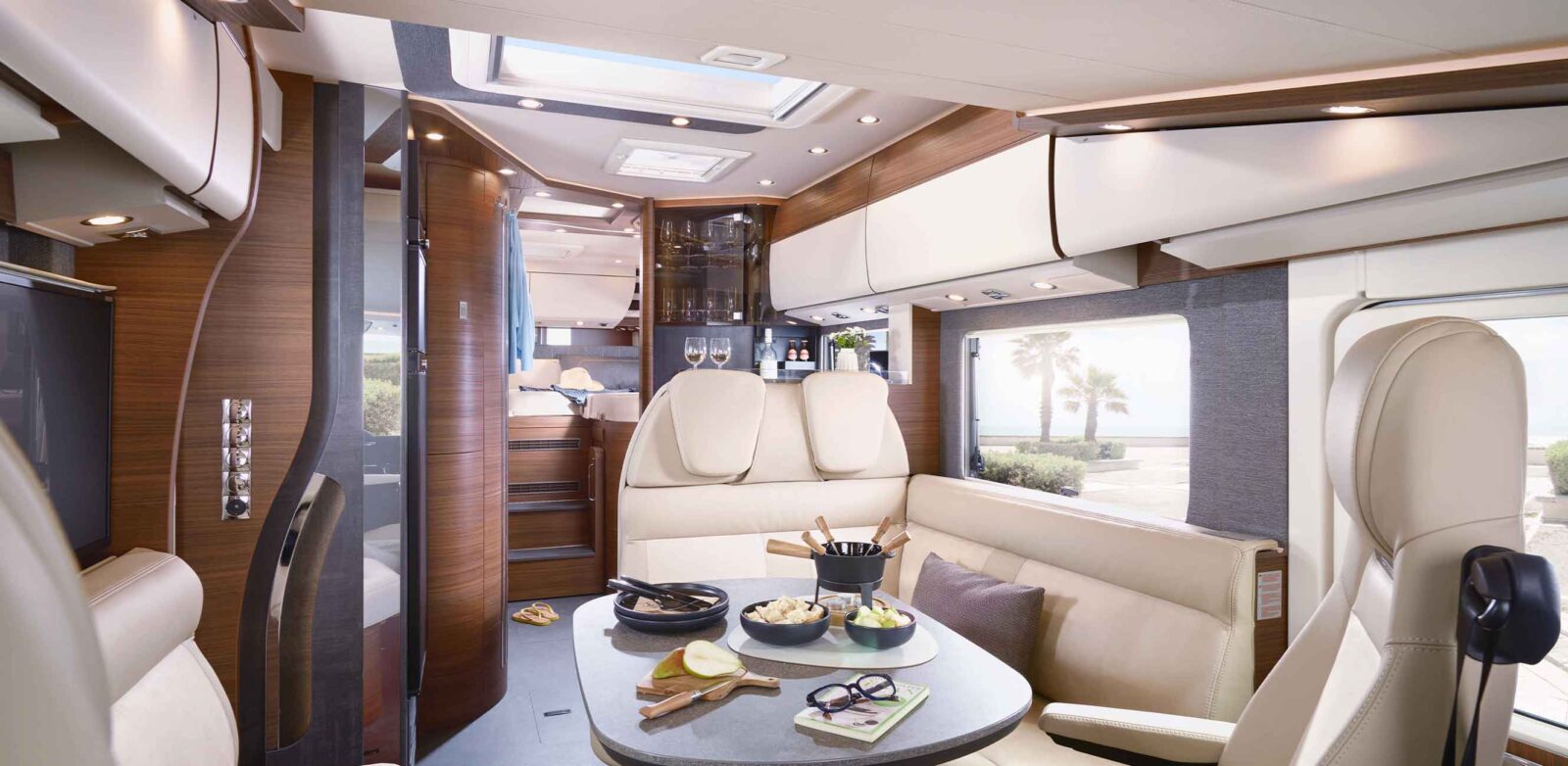 A modern motorhome interior featuring cream-colored leather seats, a dining table set with food and drinks, and wood paneling. Large windows offer a view of a sunny beach lined with palm trees—and highlight the new model year with stylish comfort.