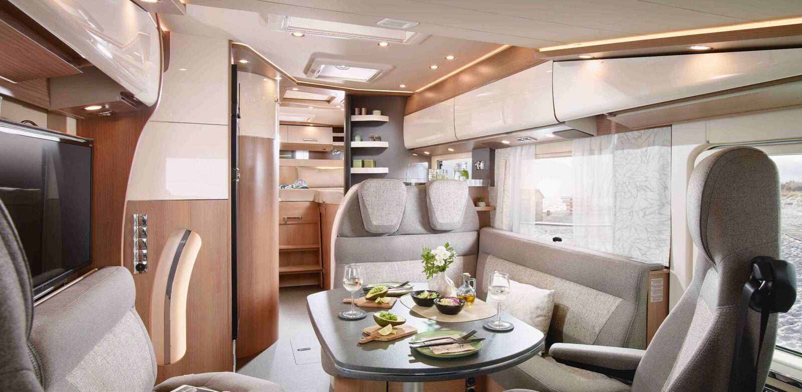 A modern, bright motorhome interior from the latest model year, featuring a set dining table with food and drinks, gray upholstered seats, overhead storage, and a compact kitchen area in the background.