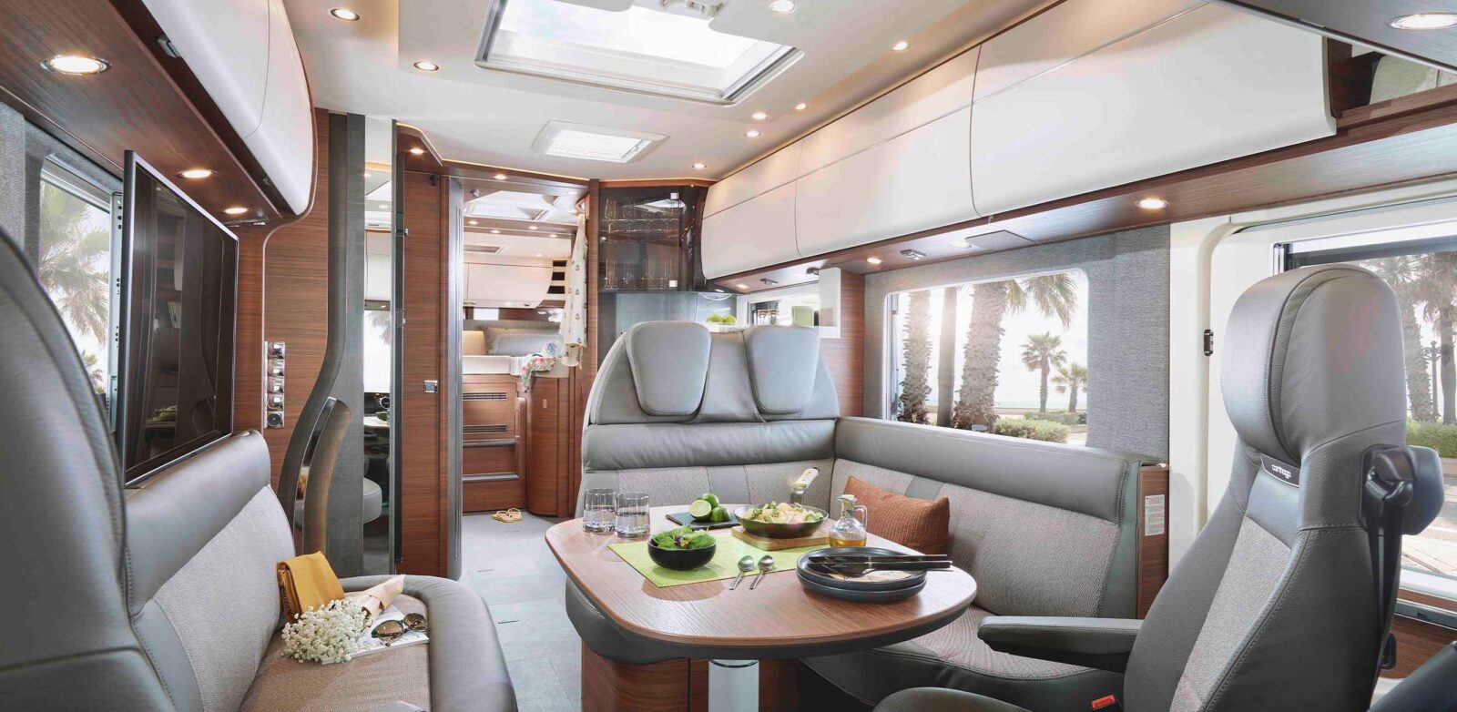 A modern motorhome interior from the latest model year, featuring a dining area for two, upholstered seats, a table set with salad and drinks, overhead storage, and large windows that let in the view of palm trees and sunlight.