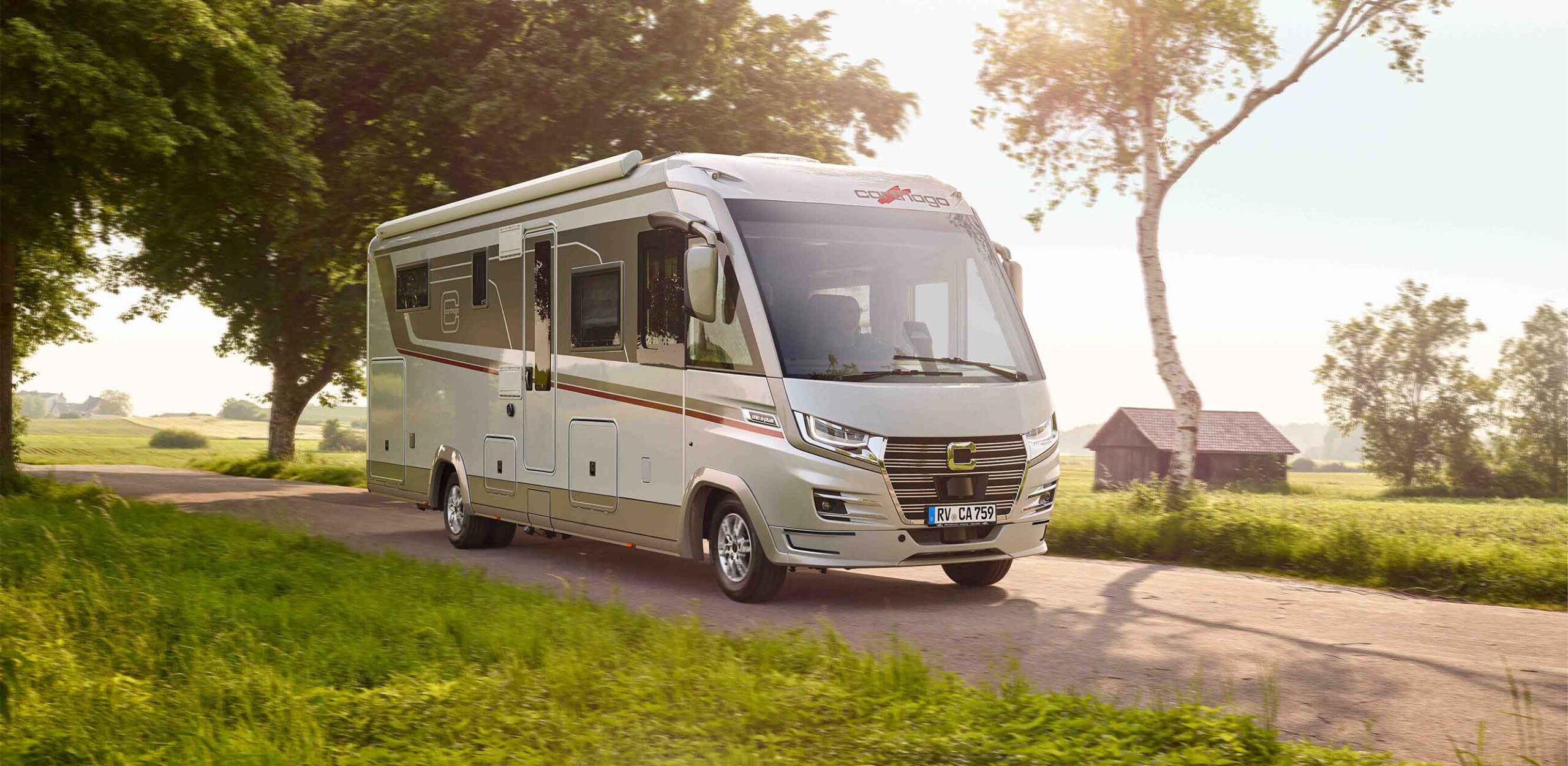 A large, modern, white motorhome with a Truma partner model drives along a country road, surrounded by green grass, trees and a wooden barn, in bright sunshine.