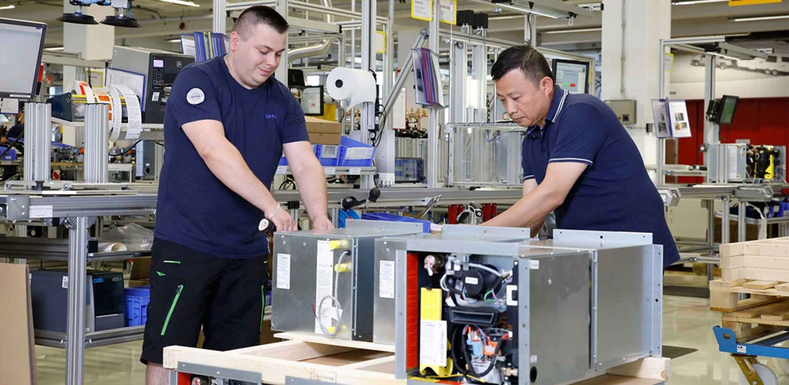 Two men assemble or test large electrical metal parts on a workbench in a factory environment, surrounded by industrial equipment and tools, during a Truma partner presentation event.
