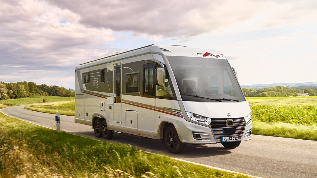 Ein modernes weißes Wohnmobil fährt auf einer Landstraße in der Nähe eines Wohnmobilstellplatzes, umgeben von grünen Feldern und Bäumen unter einem teilweise bewölkten Himmel.