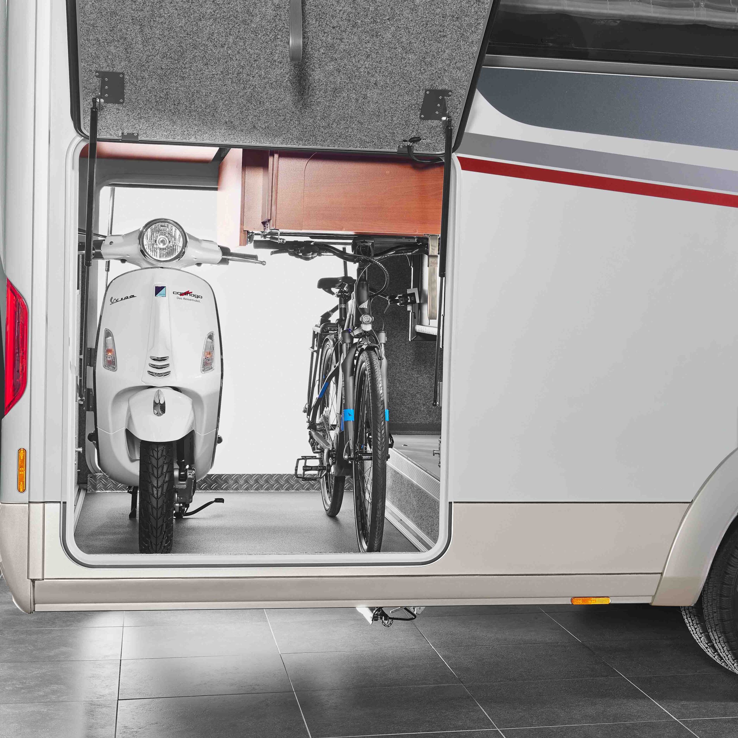 Eine Nahaufnahme eines offenen Carthago Liner for Two Wohnmobil-Staufachs, in dem ein geparkter weißer Motorroller und ein schwarzes Fahrrad nebeneinander gesichert sind.