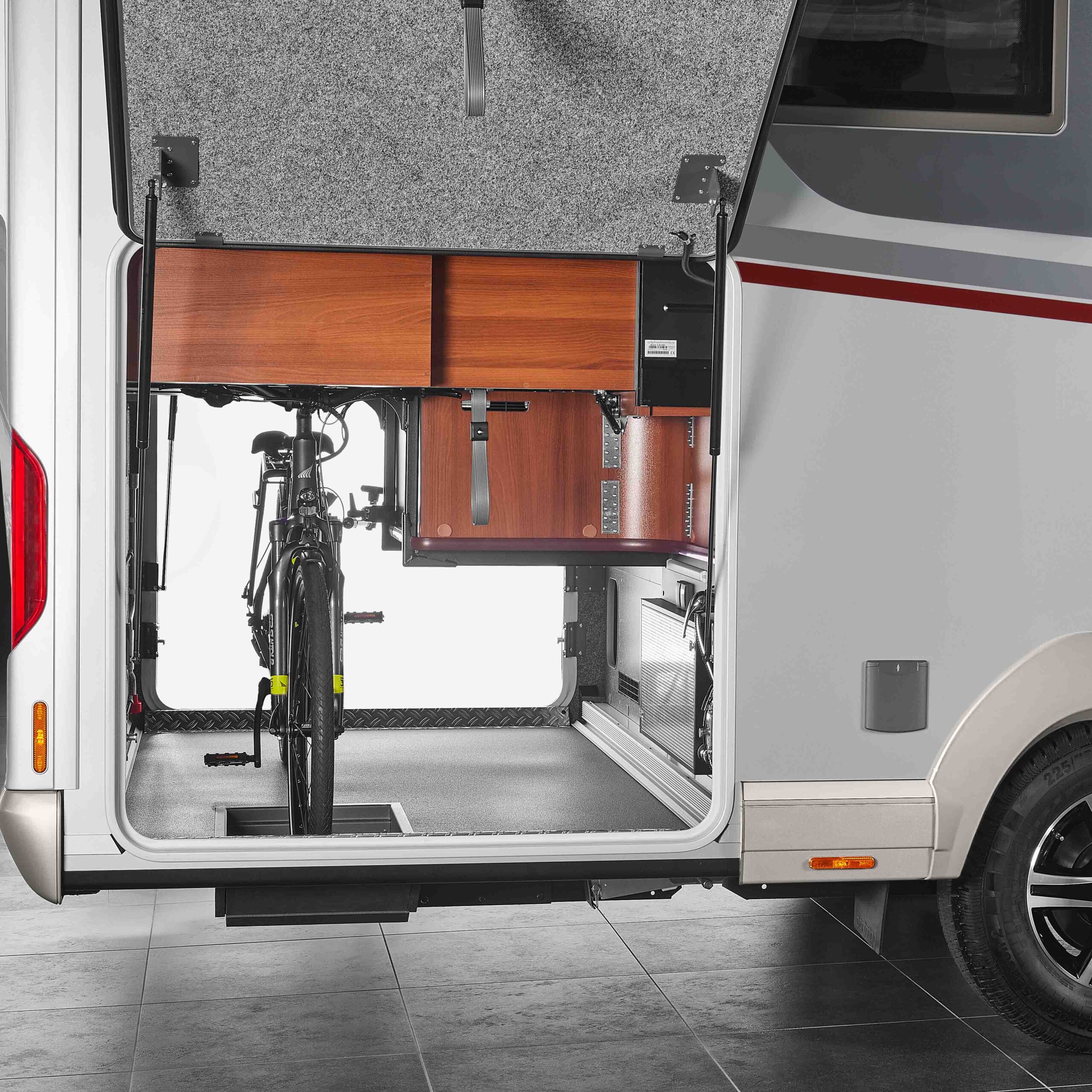 Offener hinterer Stauraum eines Carthago Liner for Two Wohnmobils mit einem darin befestigten Fahrrad, Holzschränken darüber und organisierten Stauräumen mit einer Seitentür und einem sichtbaren Hinterrad auf der rechten Seite.