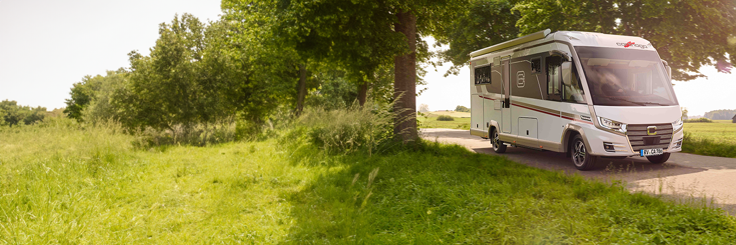 Ein Wohnmobil des Typs Carthago Liner-for-Two fährt auf einer schmalen, sonnigen Landstraße entlang, die von hohem, grünem Gras und Bäumen gesäumt wird, während im Hintergrund ein offenes Feld zu sehen ist.