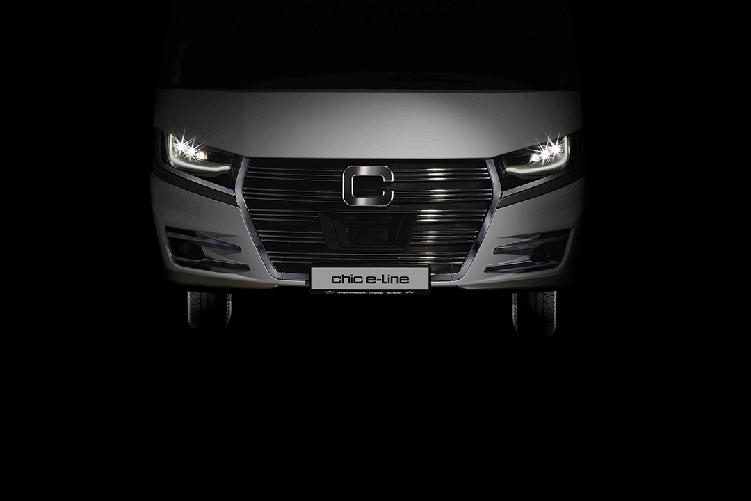 Frontansicht eines weißen Fahrzeugs mit beleuchteten Scheinwerfern, einem großen schwarzen Kühlergrill und einem Nummernschild mit der Aufschrift chic e-line vor dunklem Hintergrund - perfekt für die Carthago Wohnmobil Handelspartner- & Fahrzeugsuche.