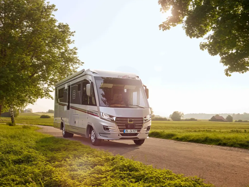Ein modernes weißes Wohnmobil vom Typ Carthago Liner for Two fährt auf einer Landstraße, umgeben von grünen Feldern und Bäumen, unter einem strahlenden, sonnigen Himmel.