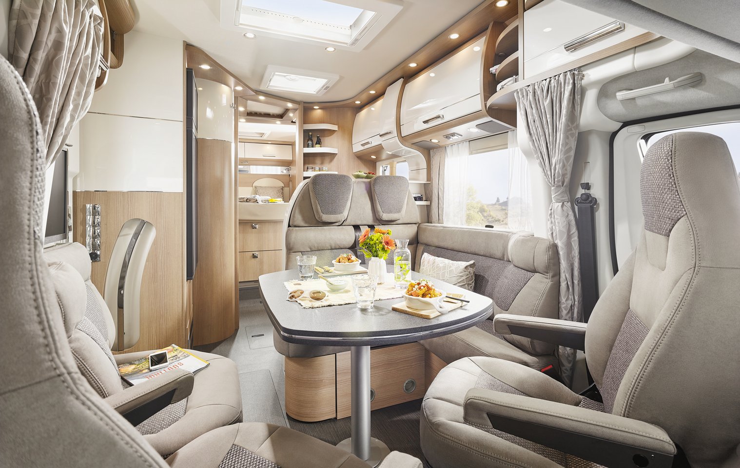 Modernes Carthago C Tourer Teilintegriertes Wohnmobil-Interieur mit beigen und hölzernen Akzenten, mit einem Esstisch mit Speisen und Getränken, gepolsterten Sitzen, Hängeschränken und großen Fenstern mit Vorhängen, die natürliches Licht hereinlassen.