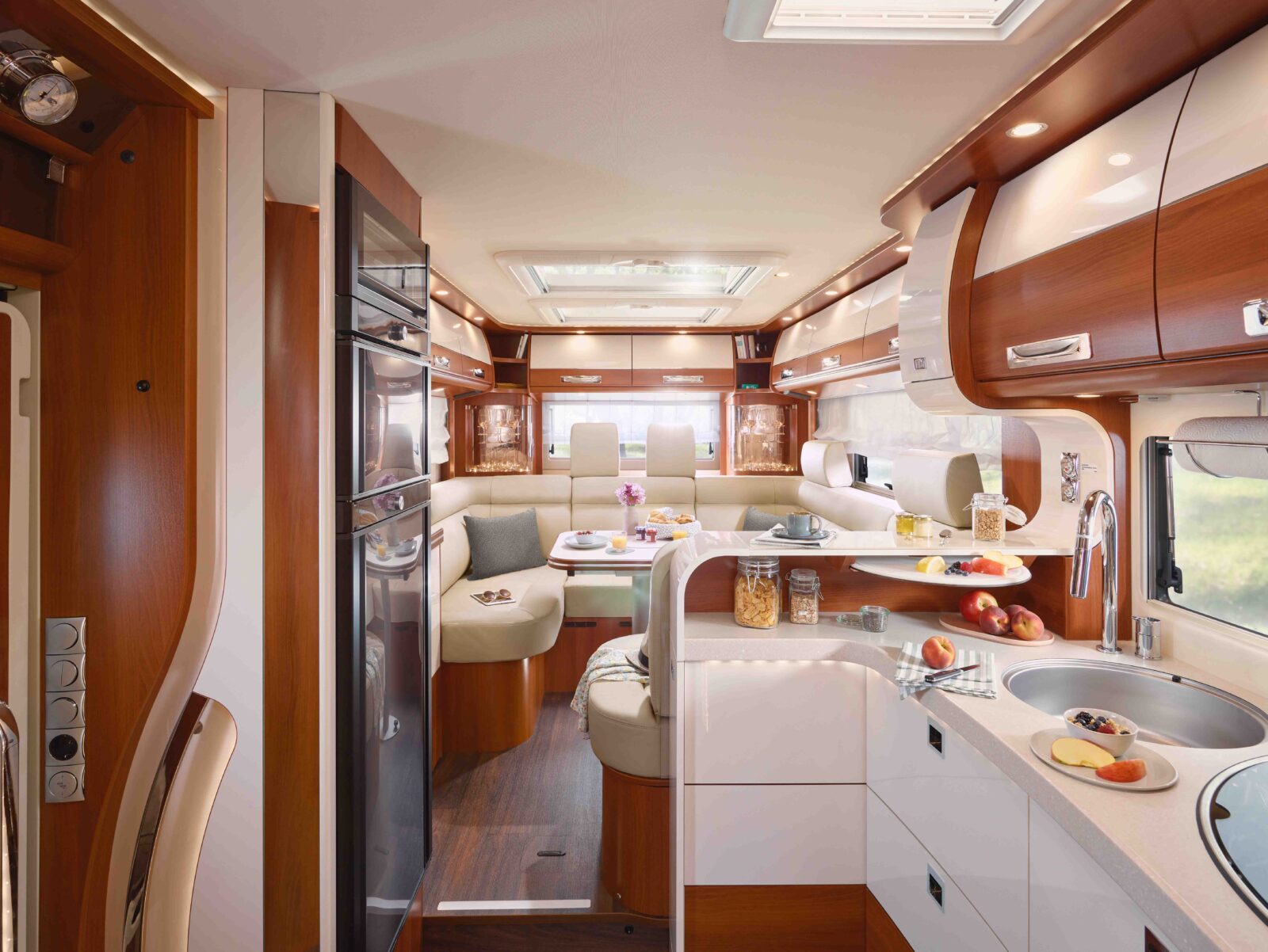 Innenraum eines modernen Carthago Liner for Two Wohnmobils mit Holzakzenten, weißen Schränken, einer Küchenzeile mit Spüle und Herd, einem geschwungenen Essbereich mit beigen Sitzgelegenheiten und Lebensmitteln auf der Theke und dem Tisch. Natürliches Licht strömt durch die Fenster.