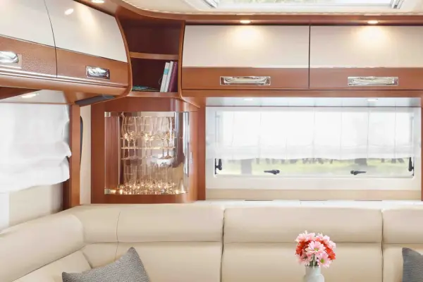 Innenraum eines modernen Wohnmobils Carthago Liner for Two mit cremefarbenen Ledersitzen, Holzschränken, einem Regal mit Gläsern, durchsichtigen weißen Vorhängen an den Fenstern und einer Vase mit rosa Blumen auf einem kleinen Tisch.