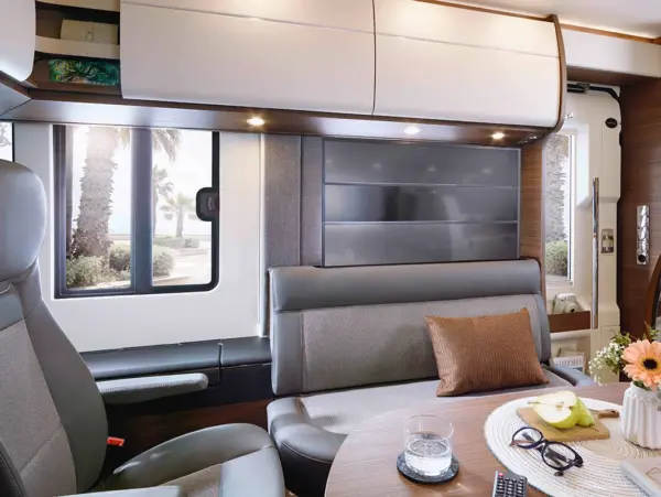 Modernes Carthago Chic C Line Wohnmobil-Interieur mit grauen Sitzen, einem Tisch mit Obst, Wasser, Blumen und einer Fernbedienung. Große Fenster geben den Blick auf Palmen und natürliches Licht frei. Überkopfschränke bieten zusätzlichen Stauraum.