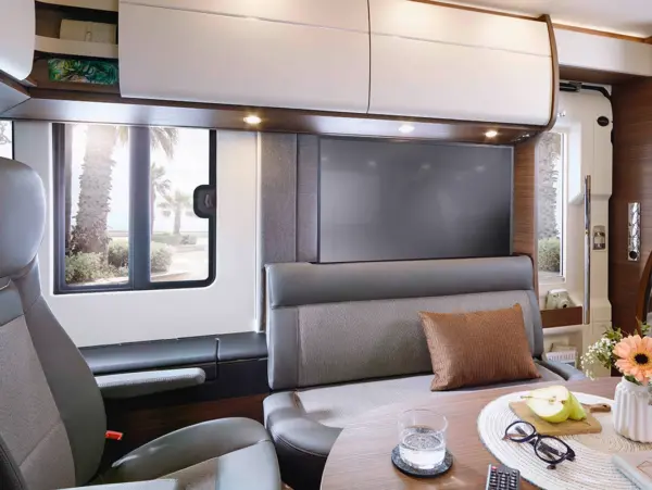 Innenraum eines modernen Carthago Chic C Line Wohnmobils mit einem grauen Sofa, einem runden Tisch mit Blumen, Obst und einem Getränk sowie großen Fenstern, die den Blick auf die Palmen im Freien freigeben.