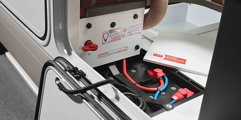 Nærbilde av batterirommet til en Carthago chic c-line T-bobil med elektriske kabler, brytere og sikkerhetsetiketter. En romdår er åpnet og kablene er koblet til batteriklemmene.