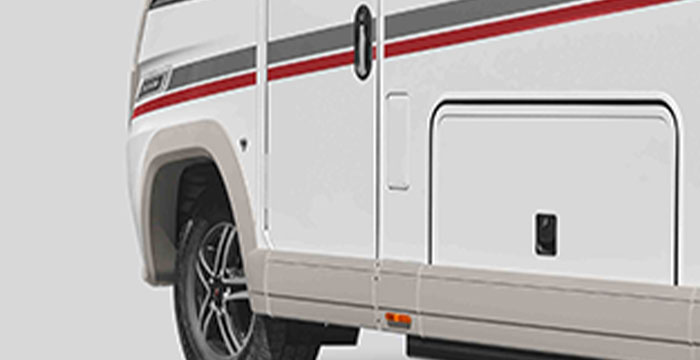 Vue partielle d’un camping-car blanc orné d’une bande rouge et grise, d’une porte de rangement et d’une partie d’une roue noire équipée d’une jante en alliage léger.