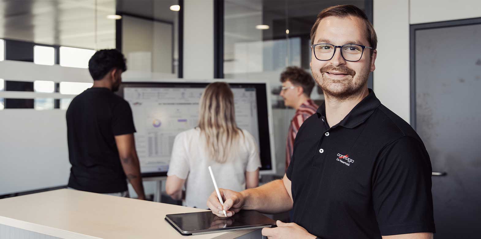 En mann med briller smiler til kameraet mens han holder en penn og et nettbrett; bak ham står tre kolleger foran en skjerm som viser diagrammer i et moderne kontormiljø.