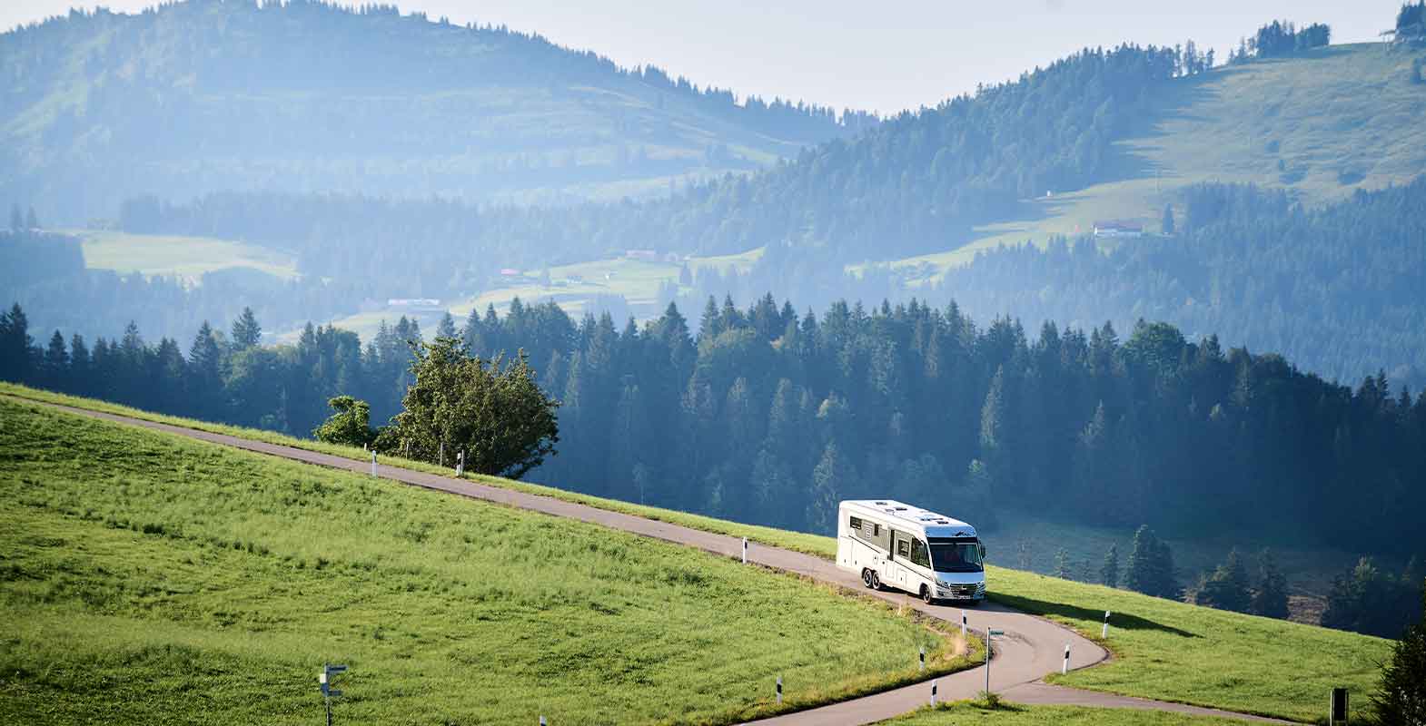Un camping-car blanc roule sur une route sinueuse à travers des collines verdoyantes, entouré d'arbres et d'un paysage montagneux aux multiples facettes, sous un ciel brumeux.