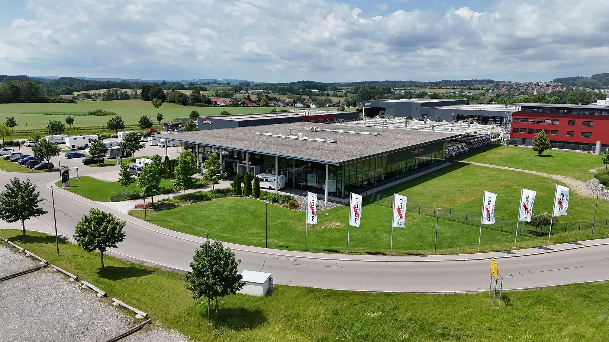 Ein modernes Industriegebäude mit großen Glasfenstern, umgeben von grünem Rasen und Fahnen, steht neben einer kurvigen Straße in einer ländlichen Gegend, in der Felder, Bäume und geparkte Autos zu sehen sind.