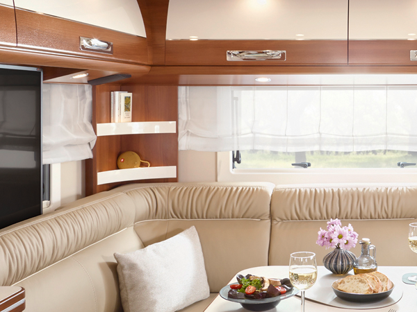 Eine gemütliche Essecke in einem Carthago Liner for Two Wohnwagen mit einem beigen Ledersofa, einem kleinen runden Tisch mit Speisen und Wein, Blumen in einer Vase und Regalen mit Büchern und einem Teddybären neben einem Fenster mit durchsichtigen Vorhängen.