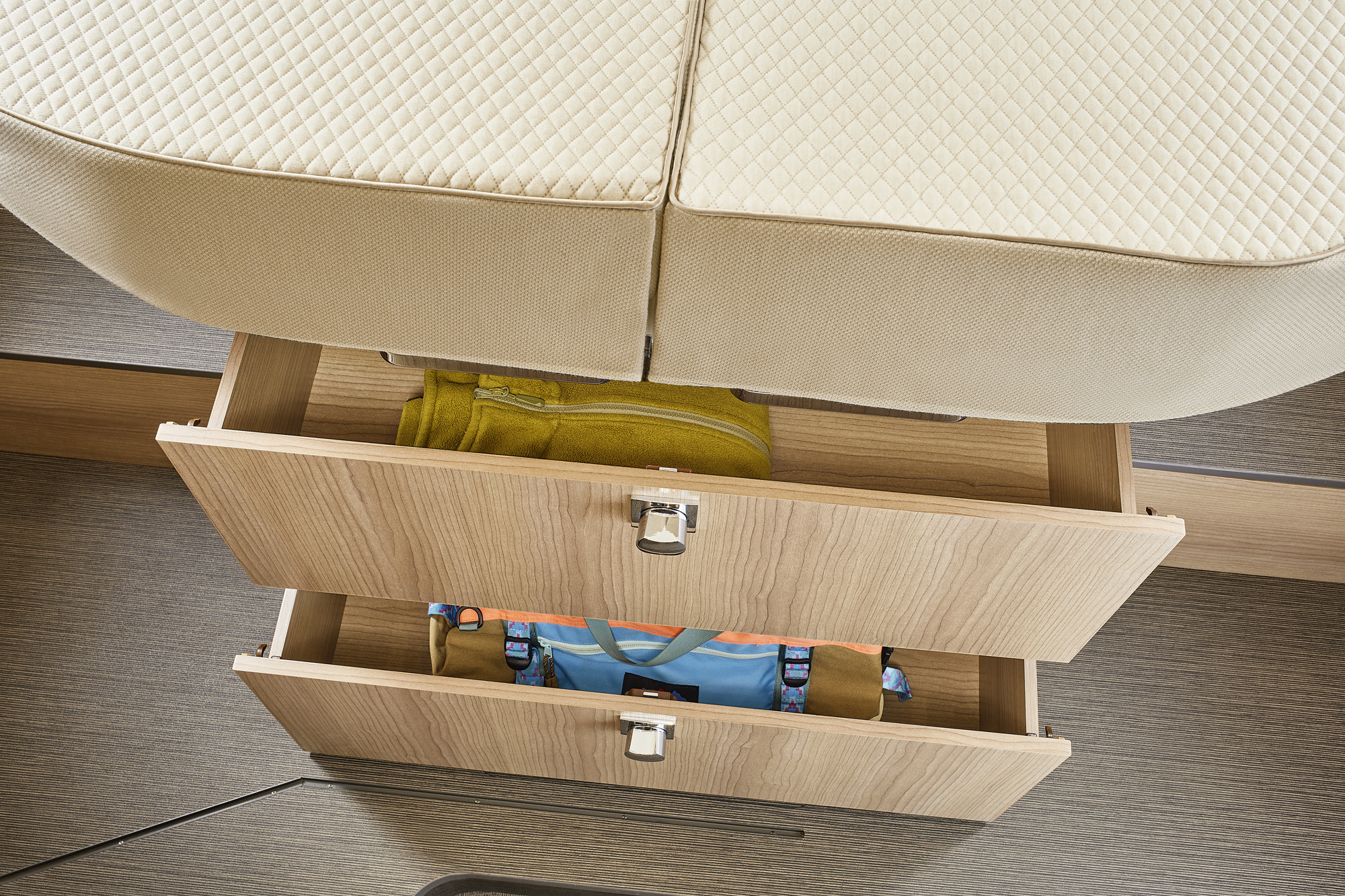 Zwei hölzerne Schubladen unter einer beigefarbenen, gesteppten Sitzbank sind teilweise geöffnet und geben den Blick auf ordentlich aufbewahrte Kleidungsstücke frei, darunter ein gelbes Handtuch und ein blaues Kleidungsstück mit bunten Trägern.