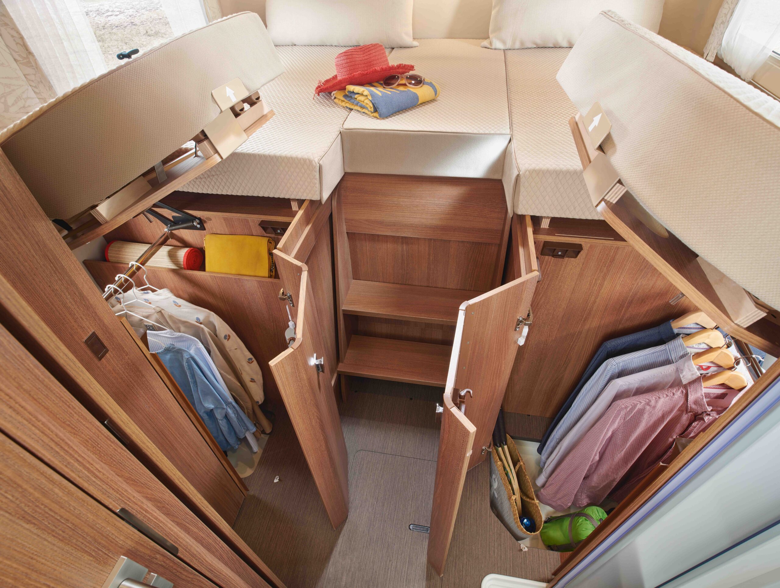 Ein kompakter, moderner Wohnmobil-Innenraum mit einem erhöhten Bett, offenen Stauschränken darunter und hängenden Kleidungsstücken in kleinen Schränken auf jeder Seite. Kleidung, Taschen und Hüte sind ordentlich im Raum verteilt.