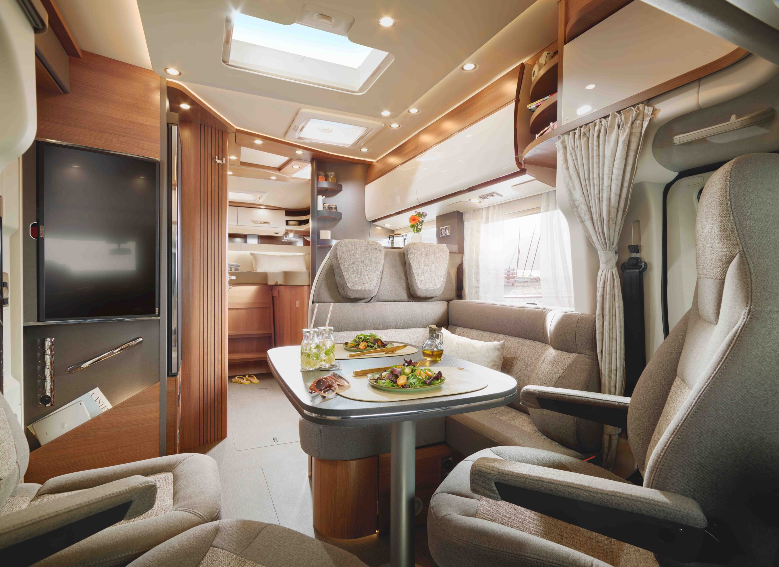 Innenraum eines modernen Wohnmobils mit beigen Sitzen, einem mit Tellern gedeckten Esstisch und einem Küchenbereich im Hintergrund. Warme Beleuchtung und große Fenster schaffen eine gemütliche, einladende Atmosphäre.