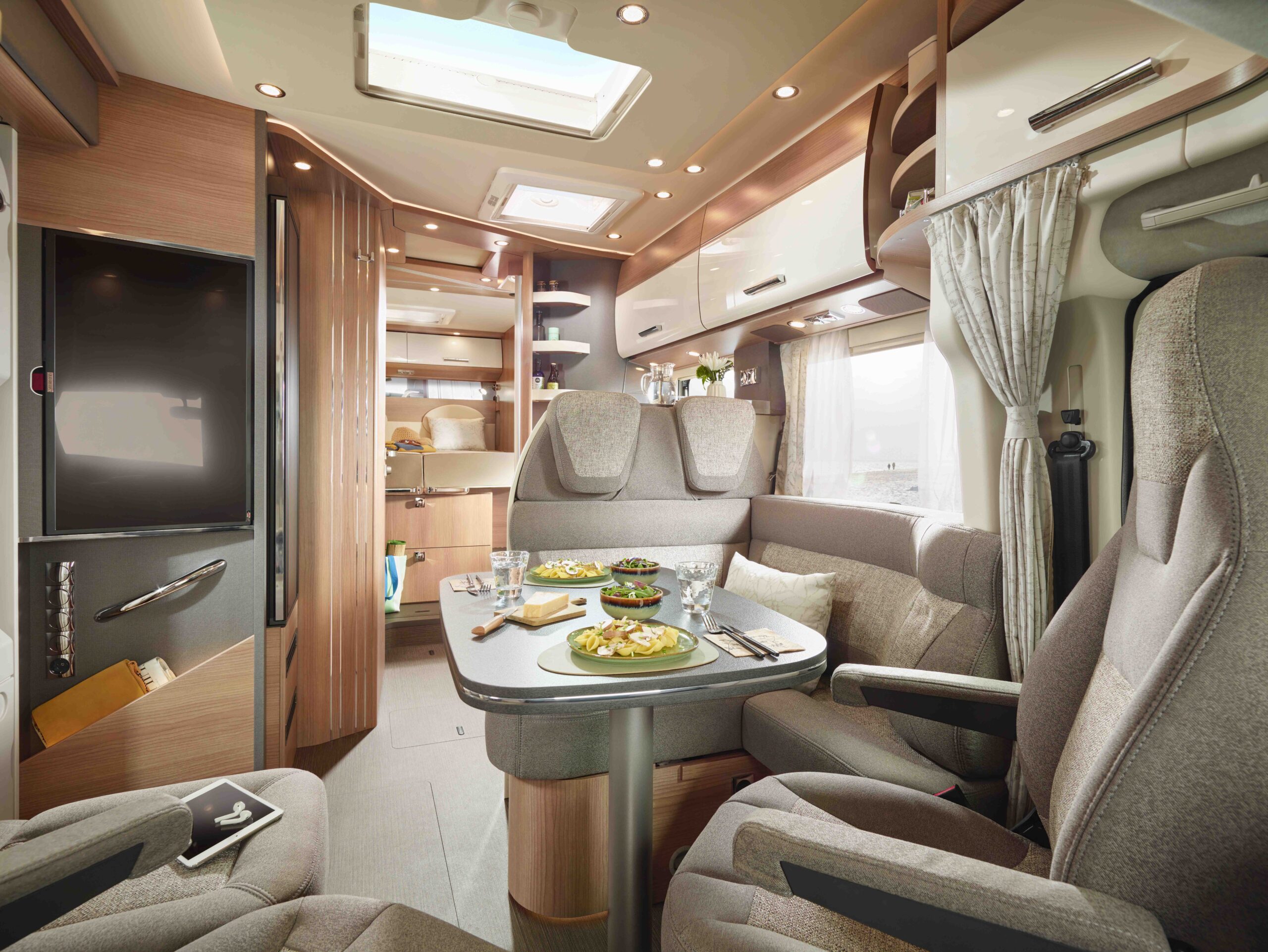 Modernes Wohnmobil-Interieur mit einem Esstisch für zwei Personen, umgeben von gepolsterten Stühlen und schlichten Schränken. Natürliches Licht fällt durch Fenster und ein Oberlicht ein und schafft eine gemütliche, einladende Atmosphäre.