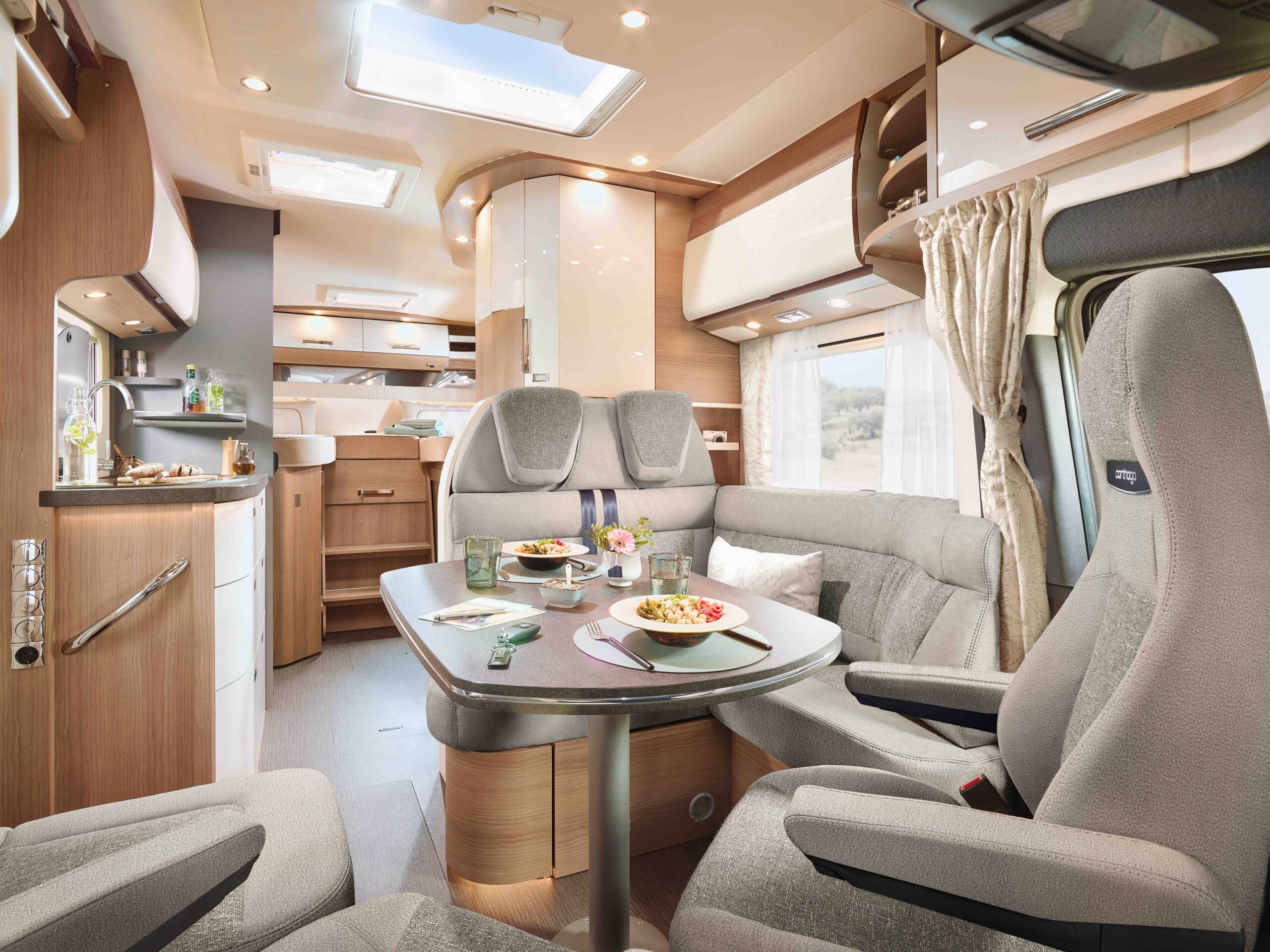 Modernes Wohnmobil-Interieur mit einem Esstisch für drei Personen mit Speisen und Getränken, umgeben von gepolsterten Sitzen, einer Küchenzeile und hellem Tageslicht, das durch große Fenster mit Vorhängen fällt.