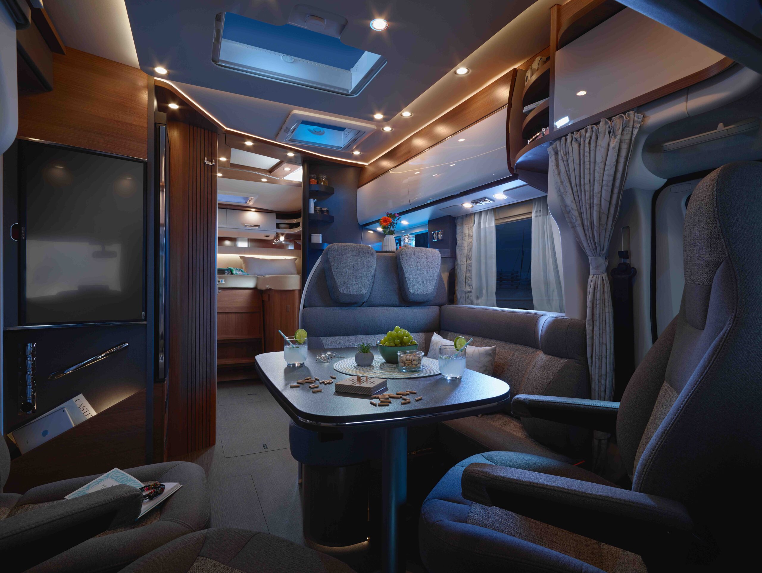 Modernes Wohnmobil-Interieur bei Nacht, mit einem Esstisch mit Getränken und Snacks, bequemen Sitzgelegenheiten, LED-Beleuchtung, Holzschränken und Blick auf die kompakte Küche.