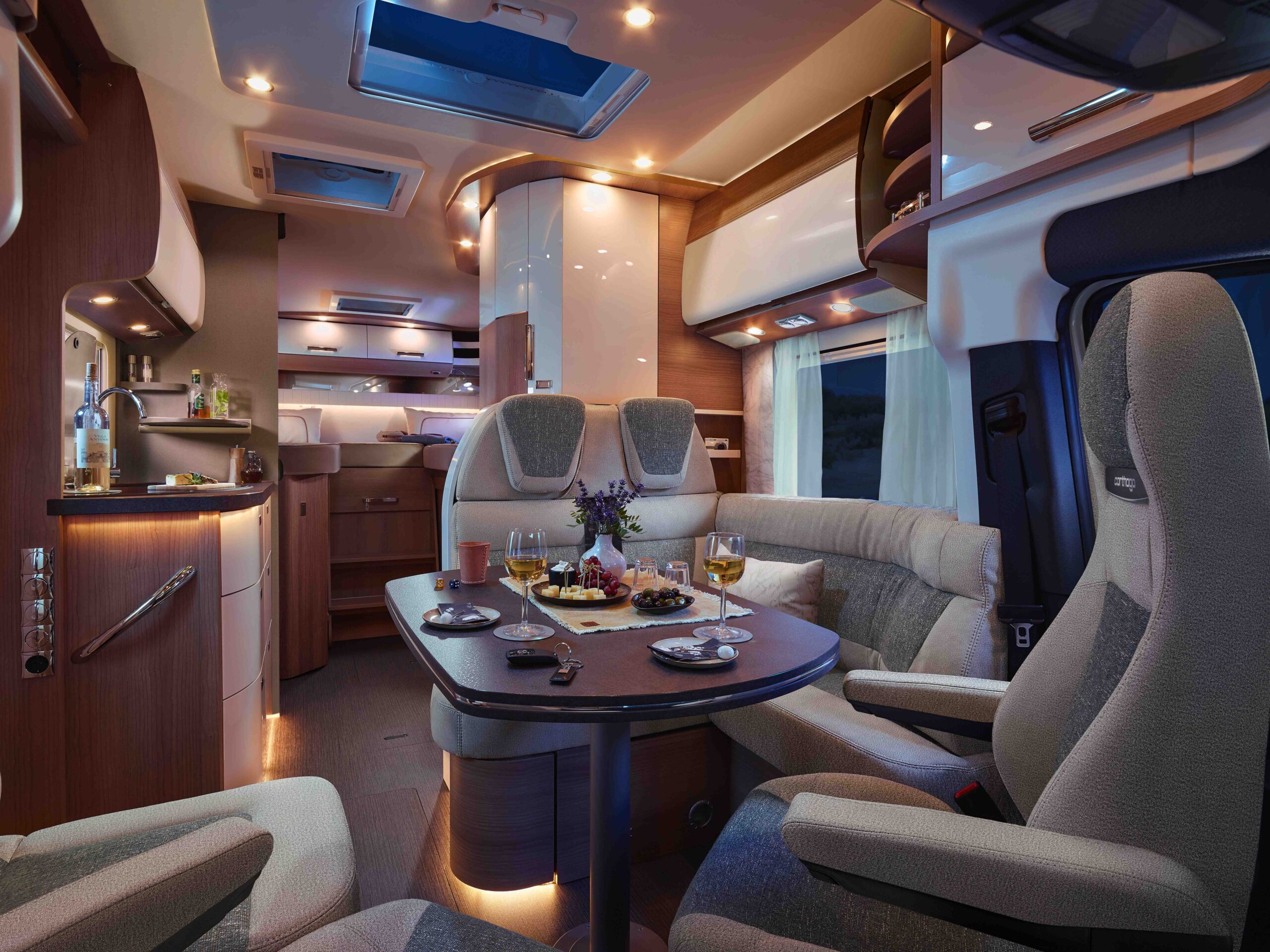 Modernes Wohnmobil-Interieur mit gedämpfter Beleuchtung und einem Esstisch mit Weingläsern, Tellern mit Speisen und Blumen. Gemütliche Sitzgelegenheiten umgeben den Tisch, und im Hintergrund sind die stilvolle Küche und der Wohnbereich zu sehen.