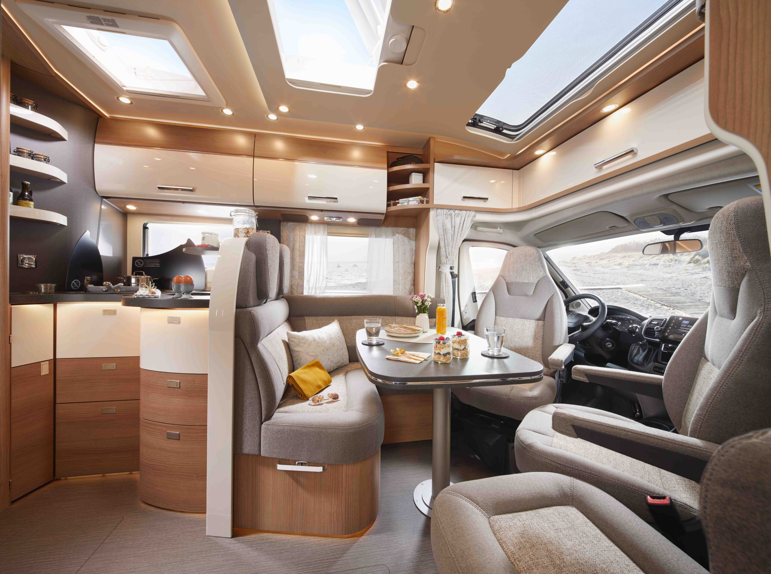 Modernes Wohnmobil-Interieur in Beige und Holz, mit einem Esstisch für zwei Personen, bequemen Sitzgelegenheiten, Stauschränken und großen Fenstern, die natürliches Licht hereinlassen.
