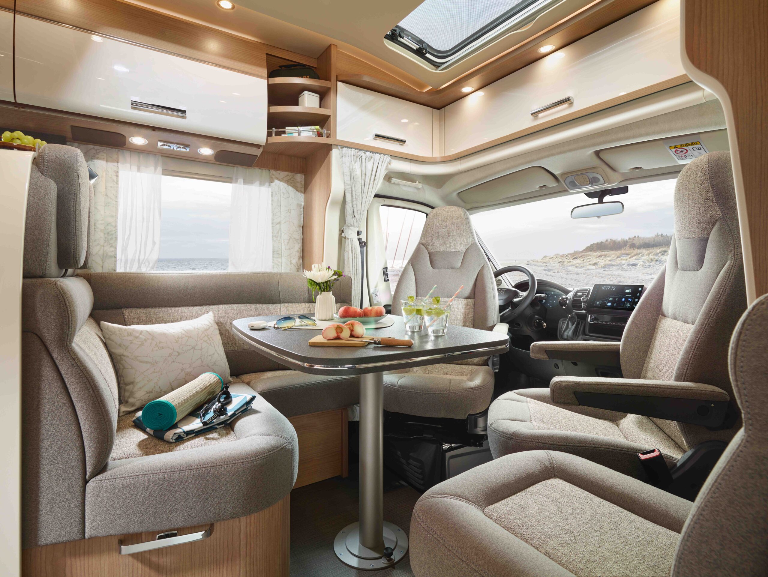Modernes Wohnmobil-Interieur mit beigen Sitzen, einem Esstisch mit Speisen und Getränken, hellem Tageslicht aus Fenstern und Oberlicht und einem malerischen Strand, der draußen zu sehen ist. Gemütlicher und gut organisierter Wohnbereich.