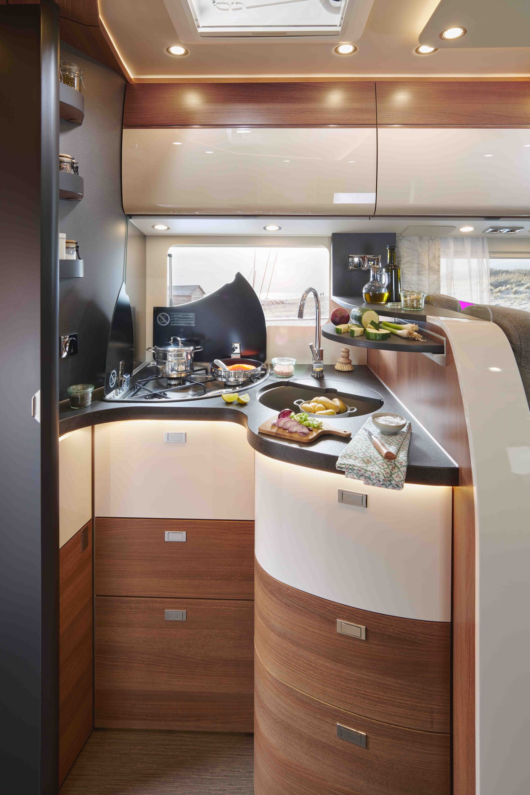 Moderne Wohnmobilküche mit geschwungenen Holzschränken, Herd mit Töpfen, Spüle und Arbeitsplatte mit Lebensmitteln und Utensilien. Regale bieten Platz für Gewürze und Wein; ein Fenster lässt natürliches Licht herein und schafft einen gemütlichen, effizienten Kochbereich.