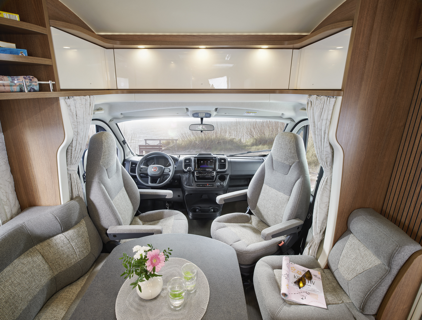 Modernes Wohnmobil-Interieur mit einer gemütlichen Sitzecke mit grau gepolsterten Sitzen, einem Tisch mit Blumen und Getränken und einem Blick auf das Armaturenbrett durch die Windschutzscheibe. Regale und natürliches Licht schaffen eine gemütliche Atmosphäre.