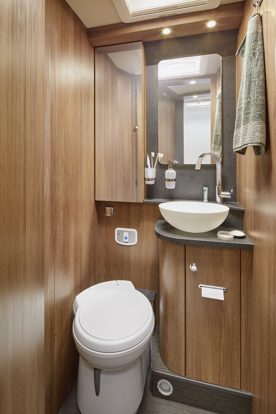 Ein kompaktes Badezimmer mit Holzschränken, einem modernen runden weißen Waschbecken auf einer schwarzen Arbeitsplatte, einem Wandspiegel, einem an einem Haken hängenden Handtuch und einer schlichten weißen Toilette. Die warme Beleuchtung erhellt den Raum.