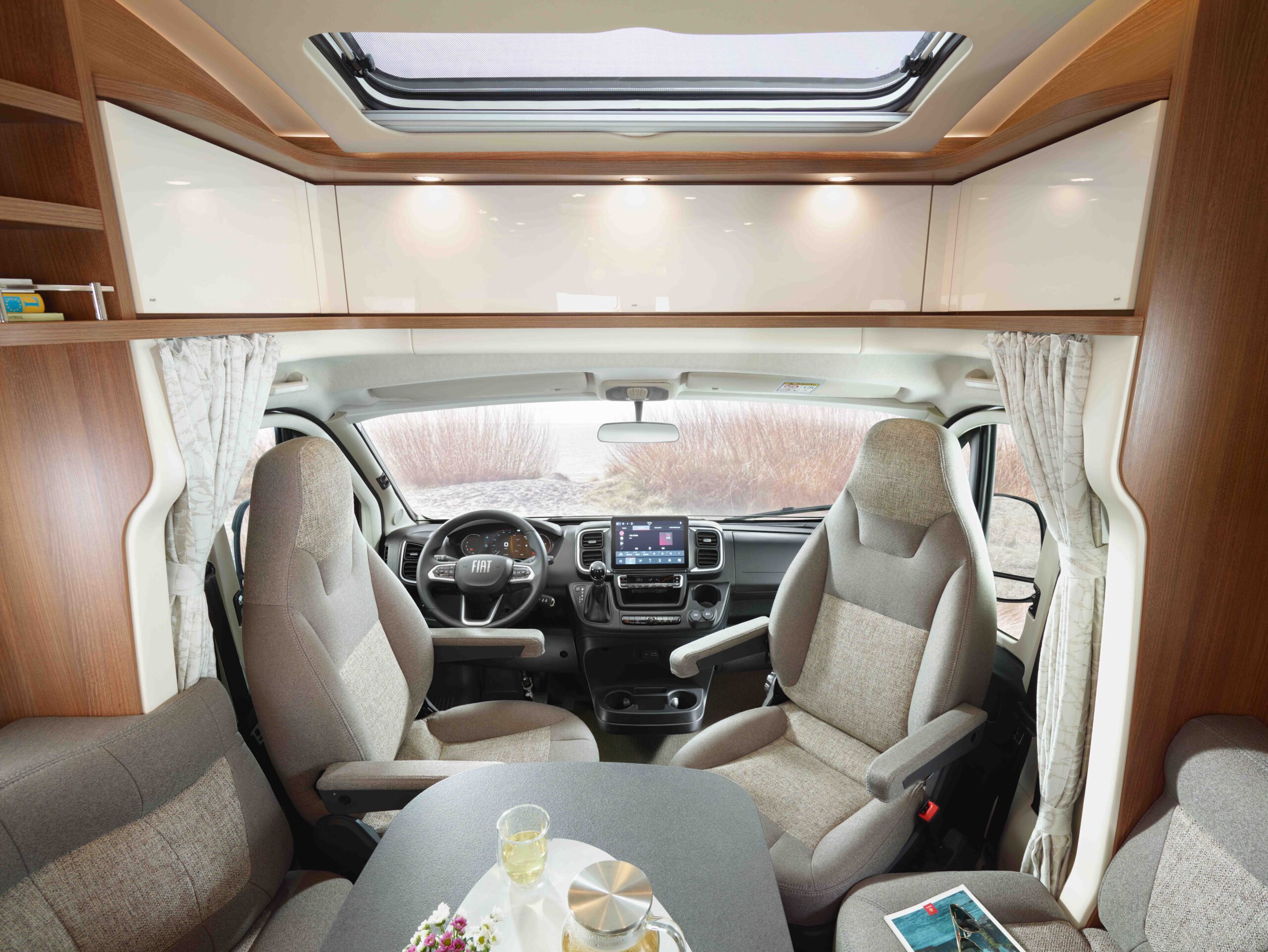 Ansicht des Innenraums eines modernen Wohnmobils mit Fahrer- und Beifahrersitz, einem Tisch mit Getränken und Snacks, hellen Holzschränken, großen Fenstern mit Vorhängen und einem Oberlicht darüber.