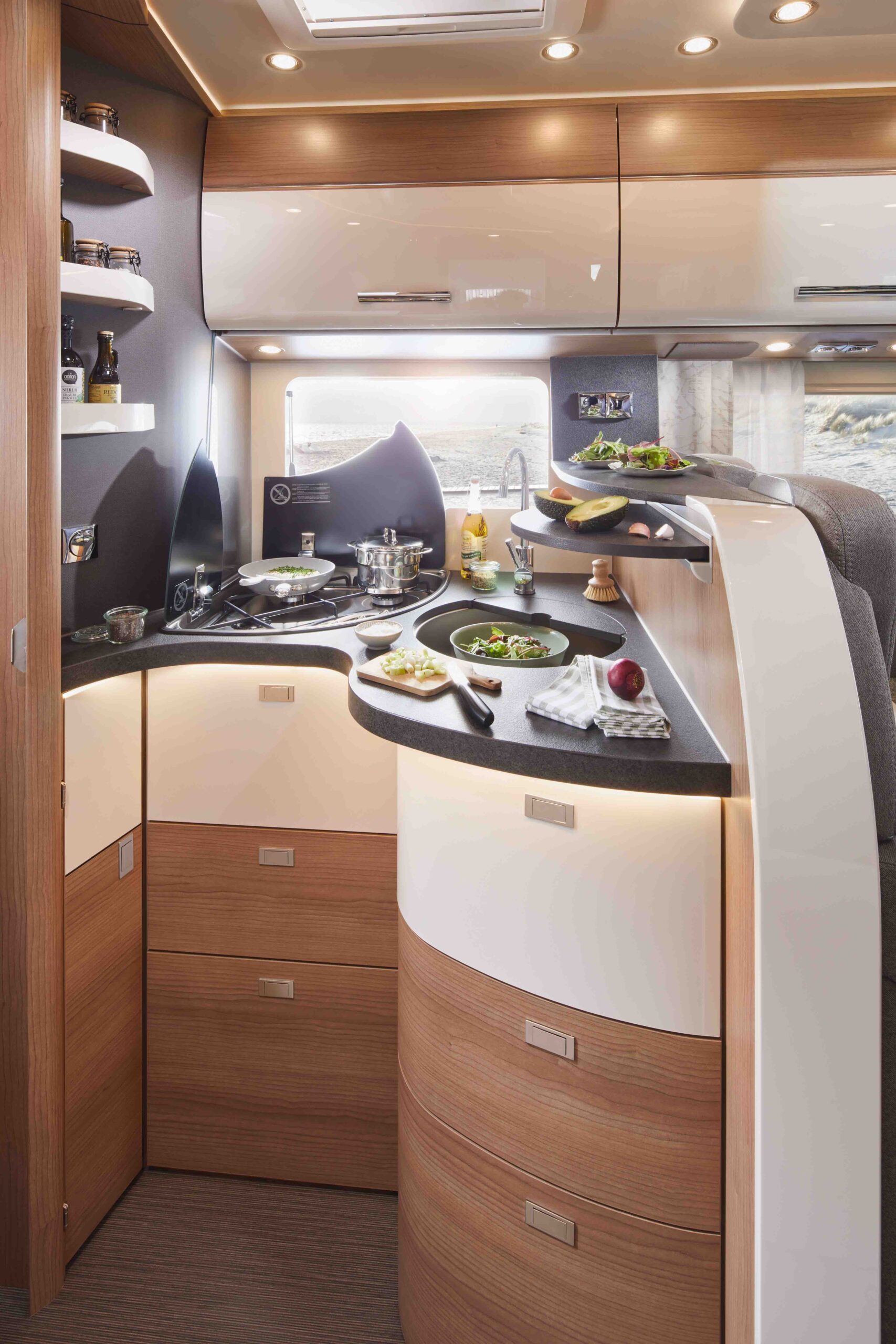Moderne Wohnmobilküche mit Holzschränken, gewölbten Arbeitsplatten, Herd, Spüle, Regalen mit Gewürzen und frischen Zutaten wie Gemüse und Kräutern auf der Theke. Helle Beleuchtung und ein Fenster sorgen für eine gemütliche Atmosphäre.